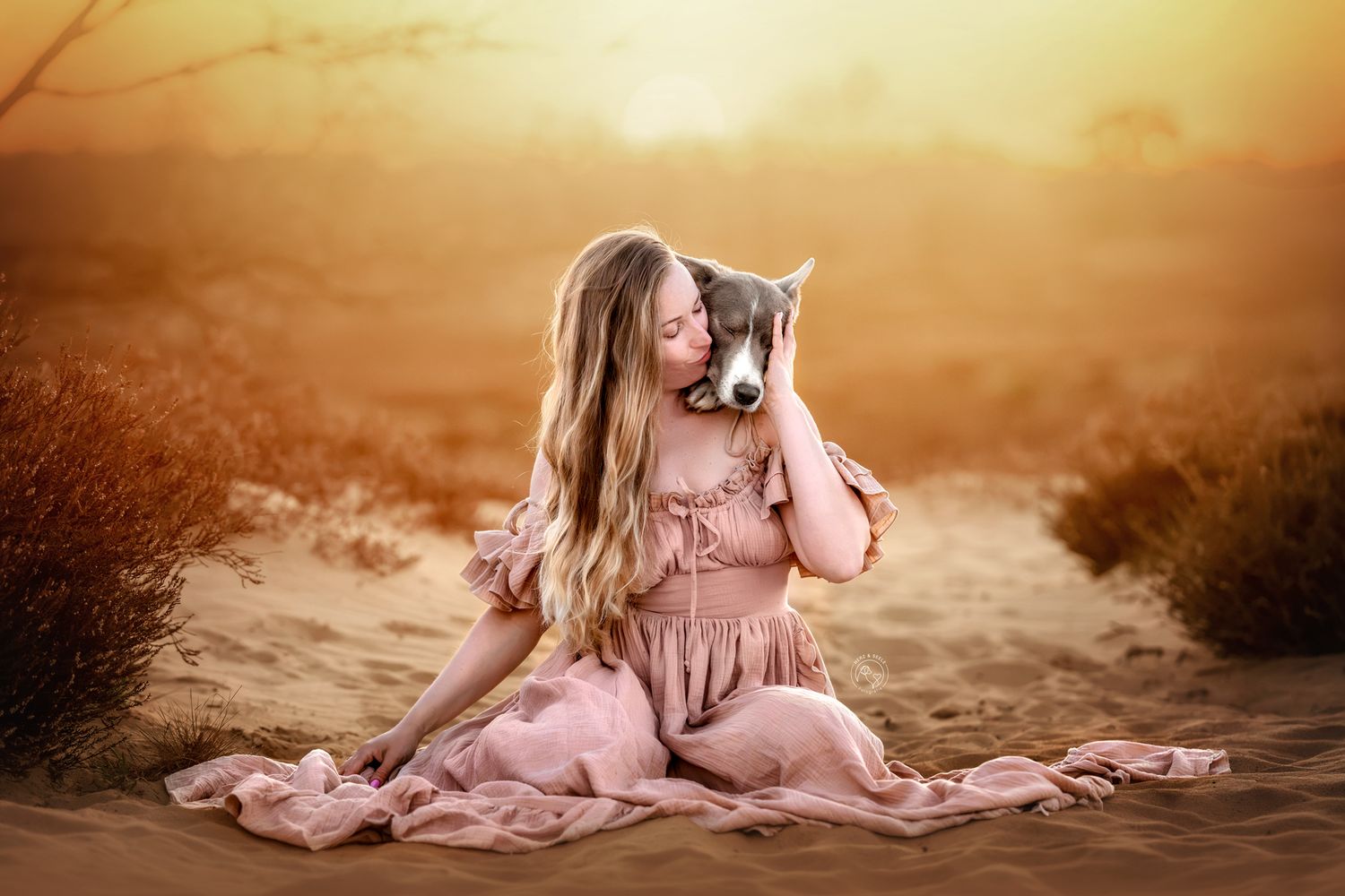 Girl in pink dress sitting on sand dunes at sunset, lovingly holding a puppy close to her face.