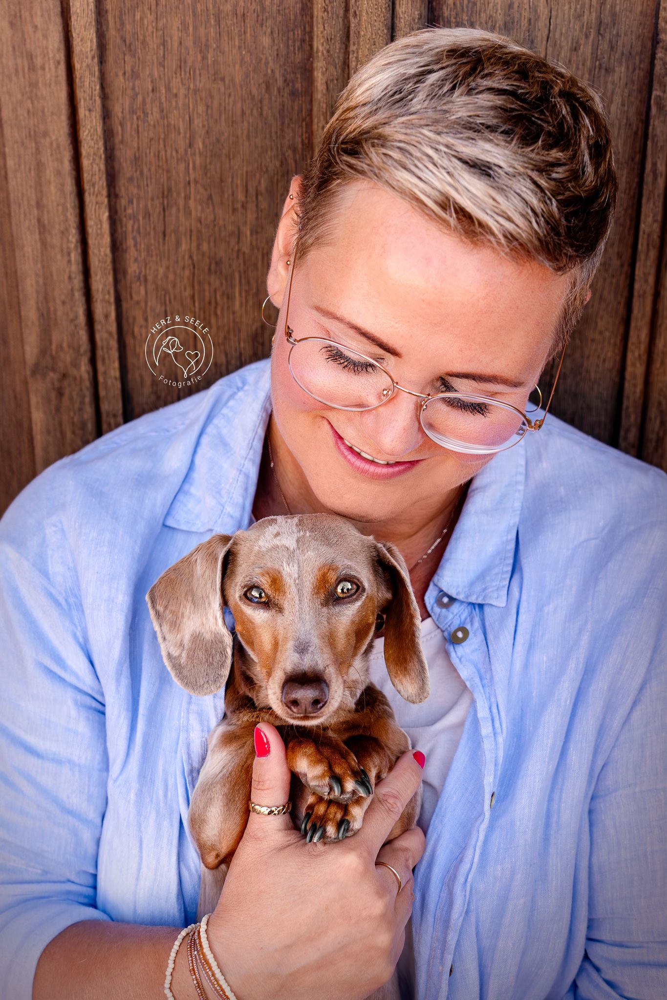 A smiling blonde woman with glasses holds an adorable dachshund puppy against a wooden wall.