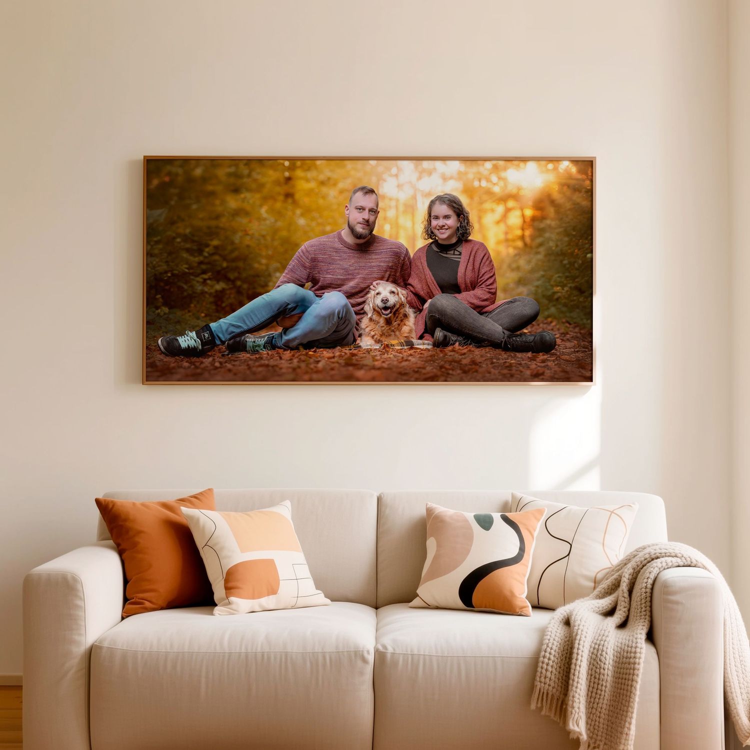 A framed panoramic photo of a couple and their golden retriever dog in autumn forest hangs above a white sofa.