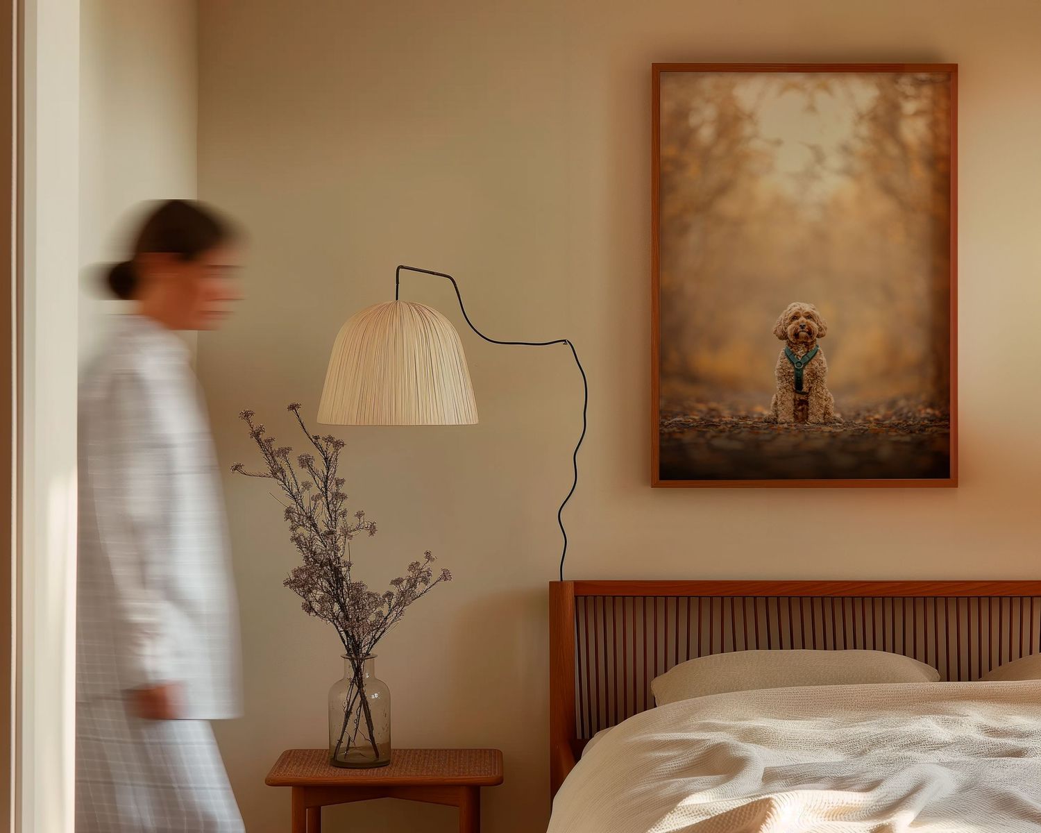 Blurred figure in white robe walks past framed dog portrait above bedside table with lamp in serene bedroom.