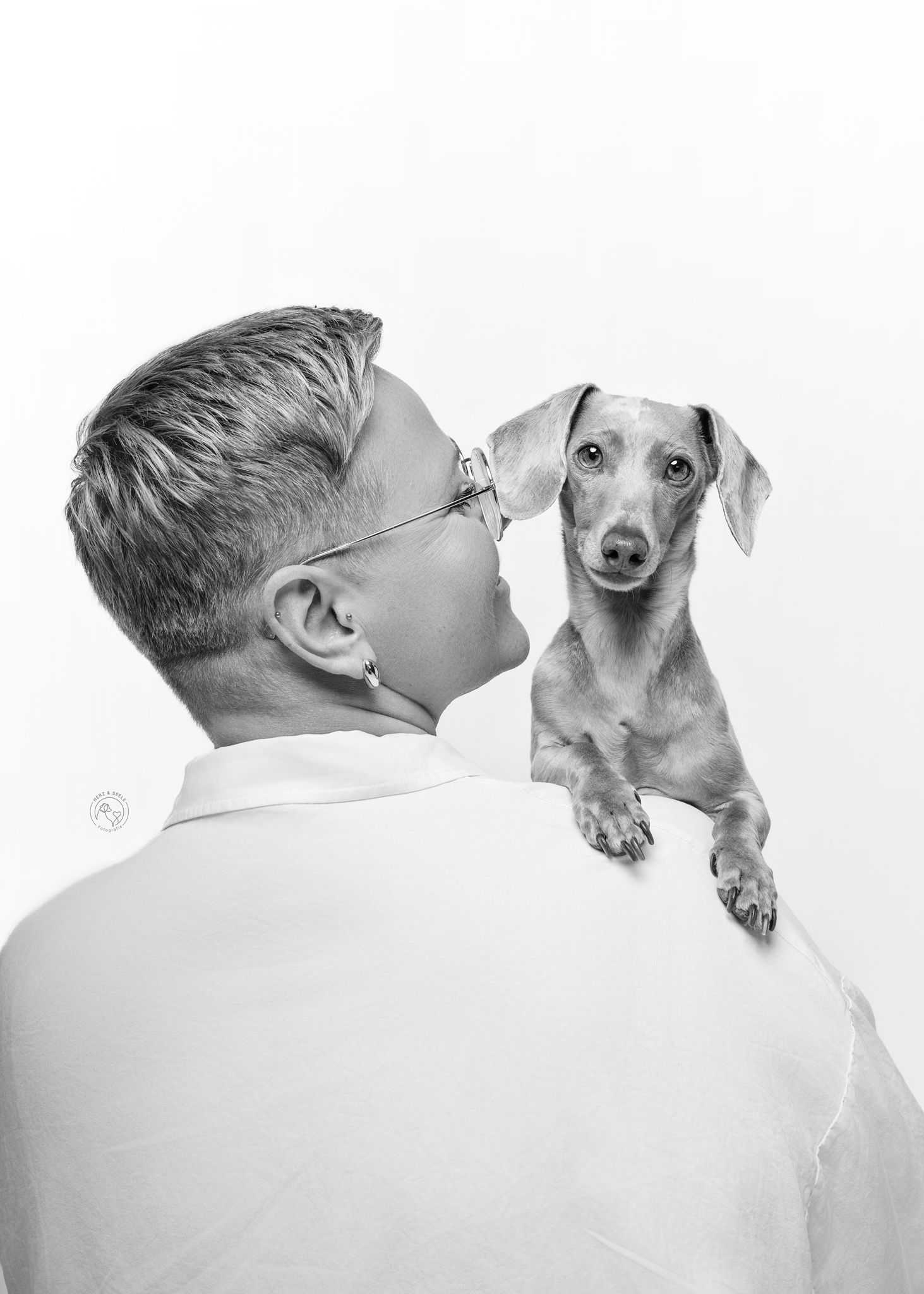 Black and white portrait of person kissing a dachshund dog over their shoulder.
