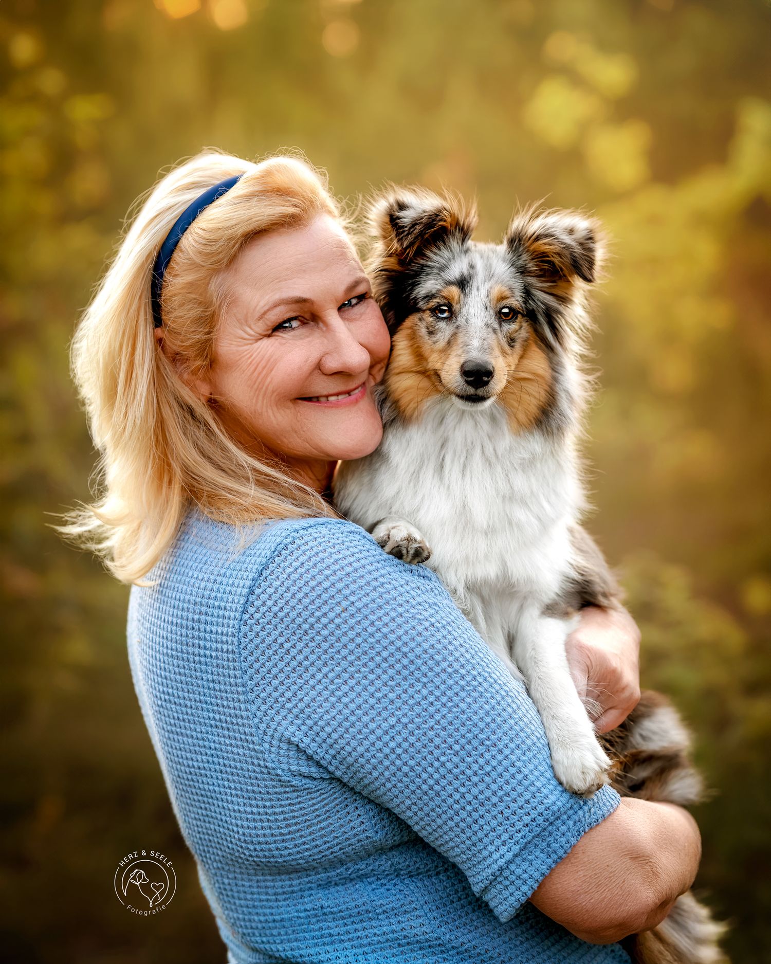 A smiling blonde woman in blue holds a small merle Sheltie puppy in golden autumn light.