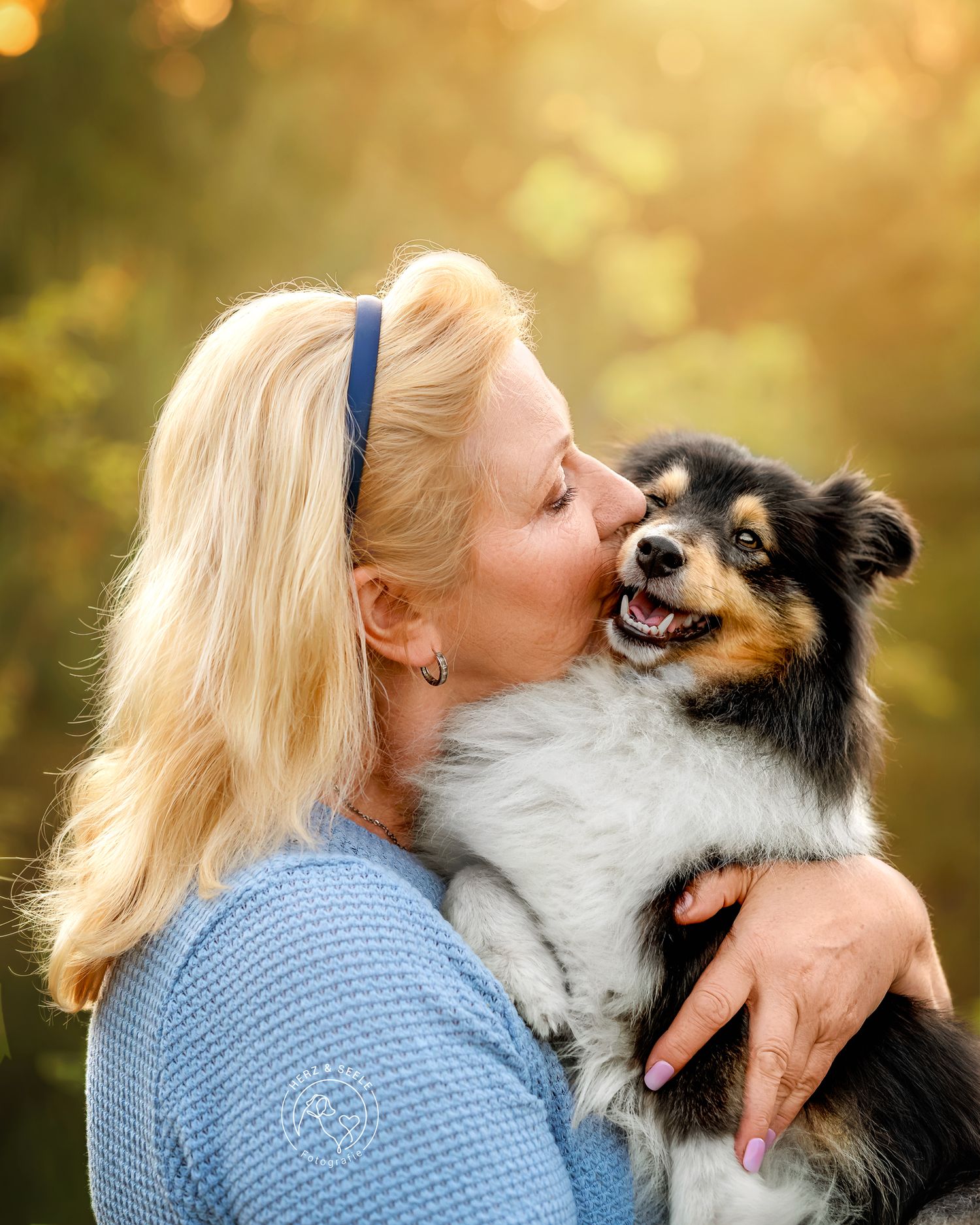 A blonde woman in blue kisses her happy tricolor Sheltie dog outdoors in golden light.