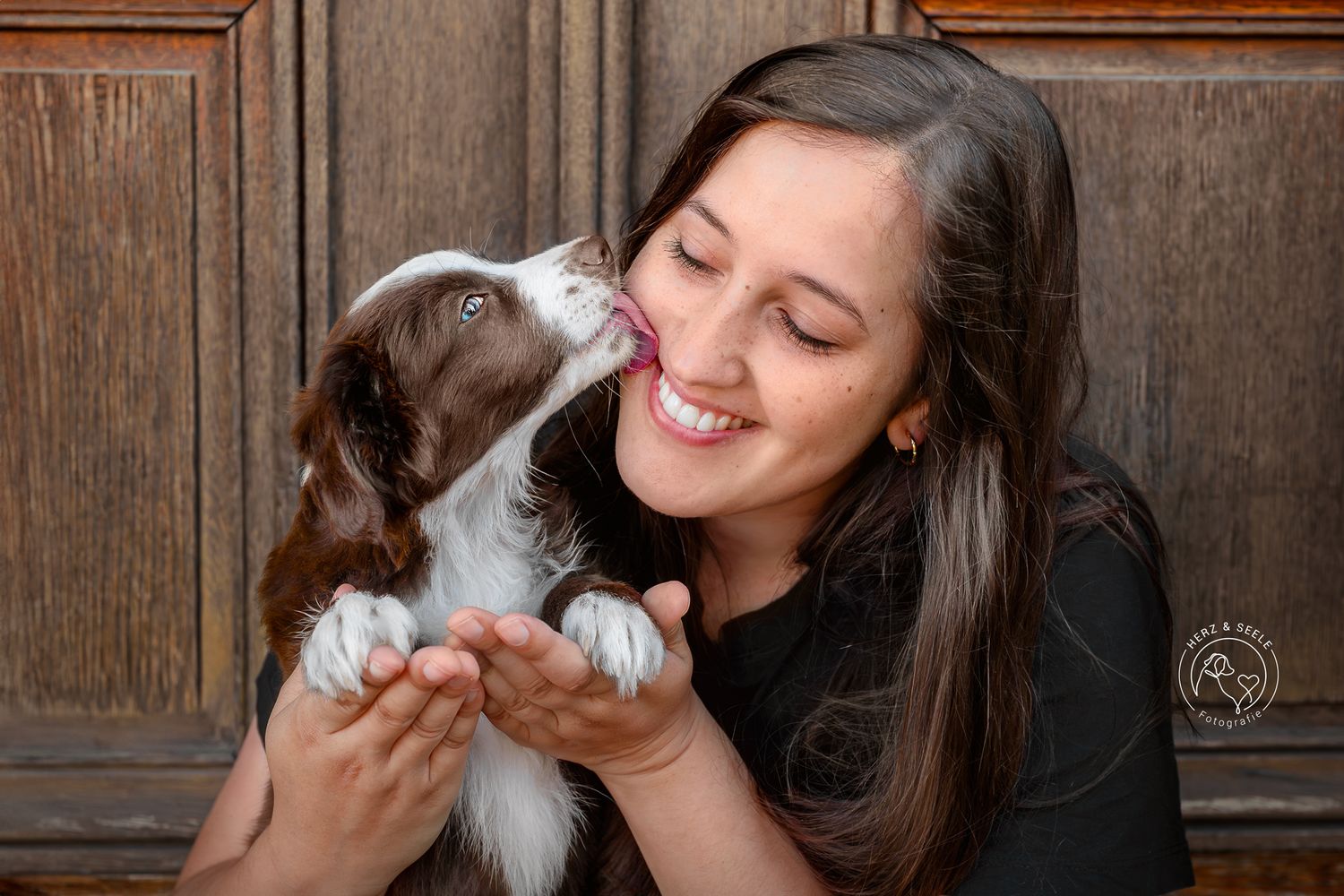 A laughing dark-haired woman gets a playful kiss from a Border Collie puppy.
