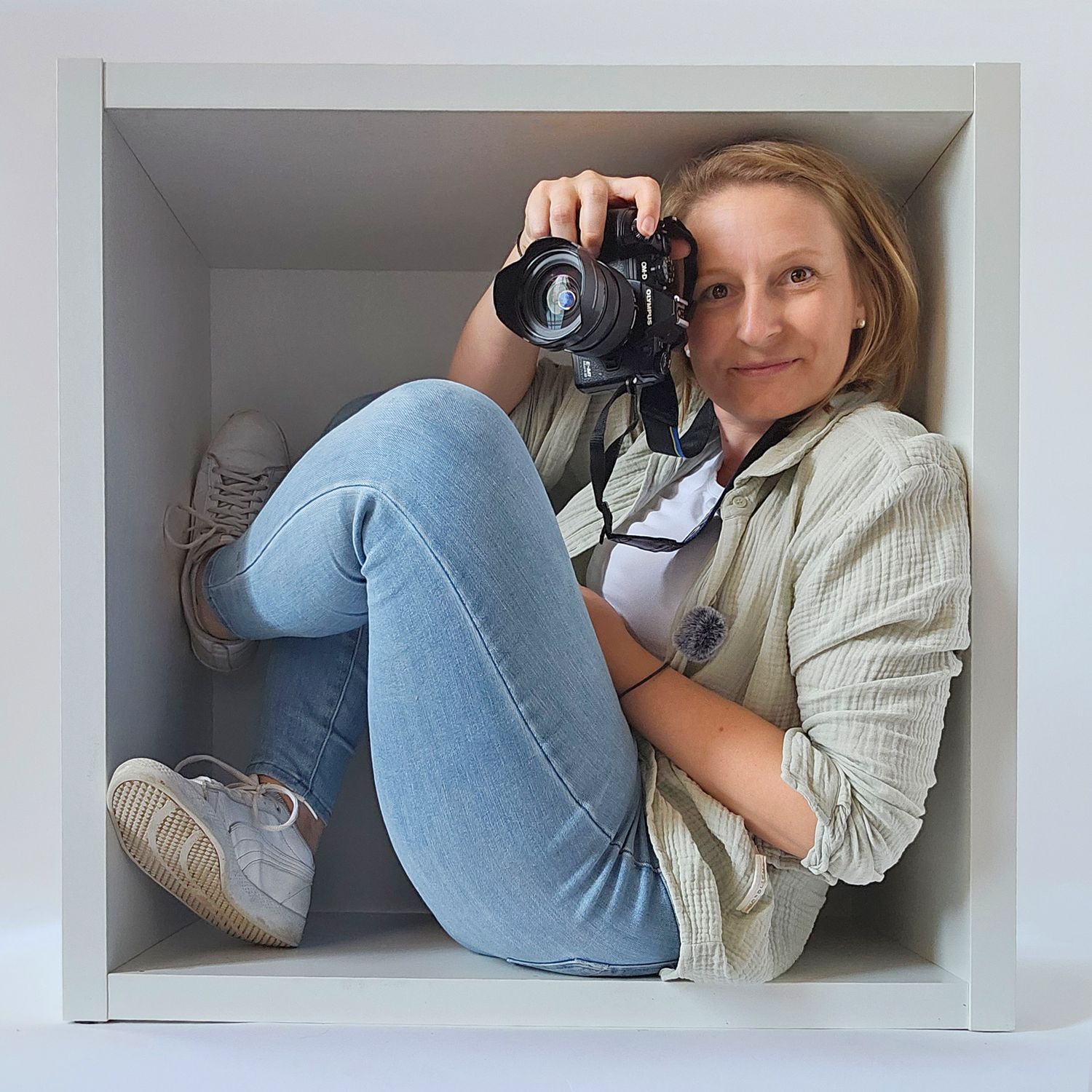 A smiling woman sits inside a white cube shelf holding a DSLR camera, wearing jeans and a beige shirt.