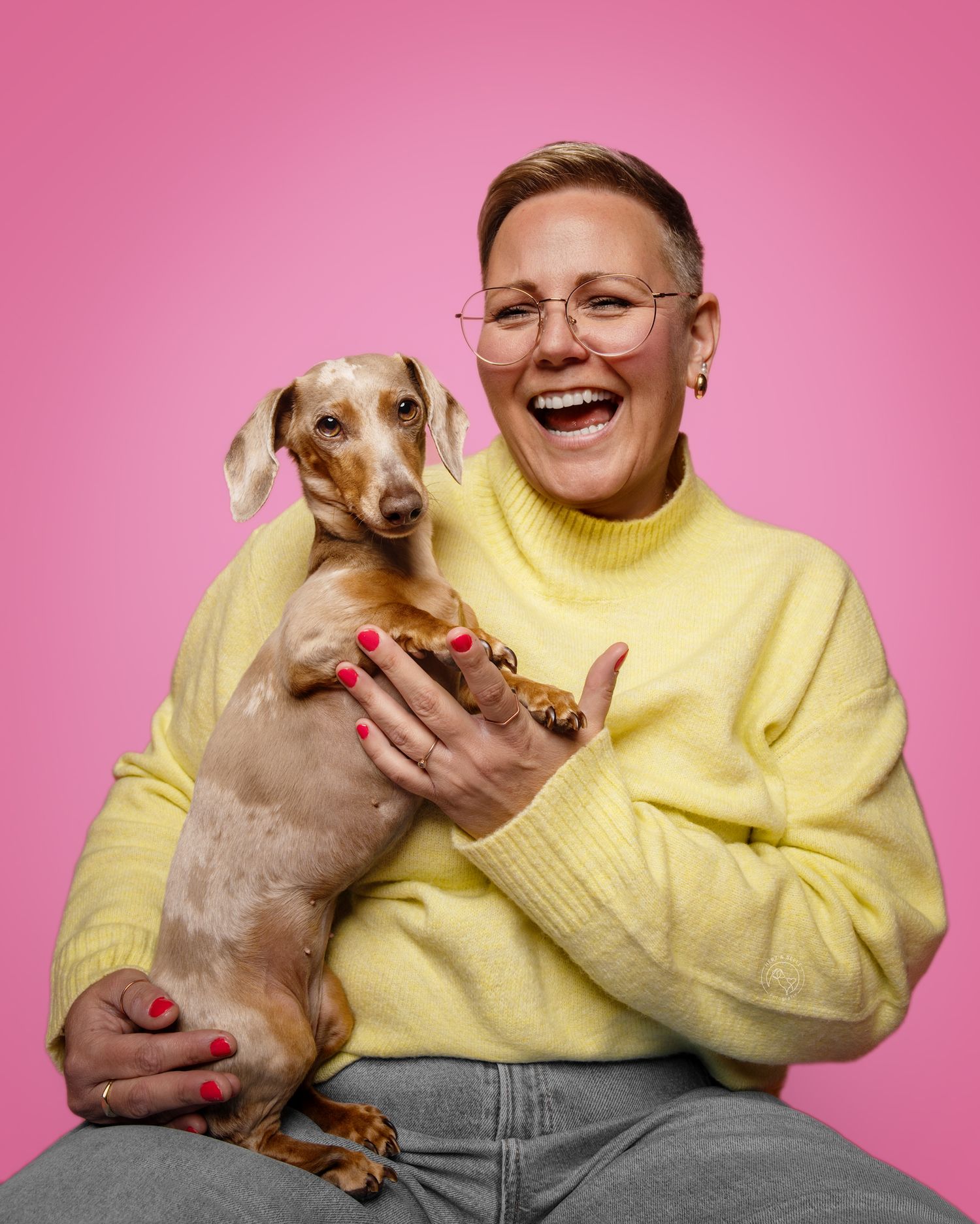 A laughing woman in a yellow turtleneck sweater holds a dachshund dog against a bright pink background.