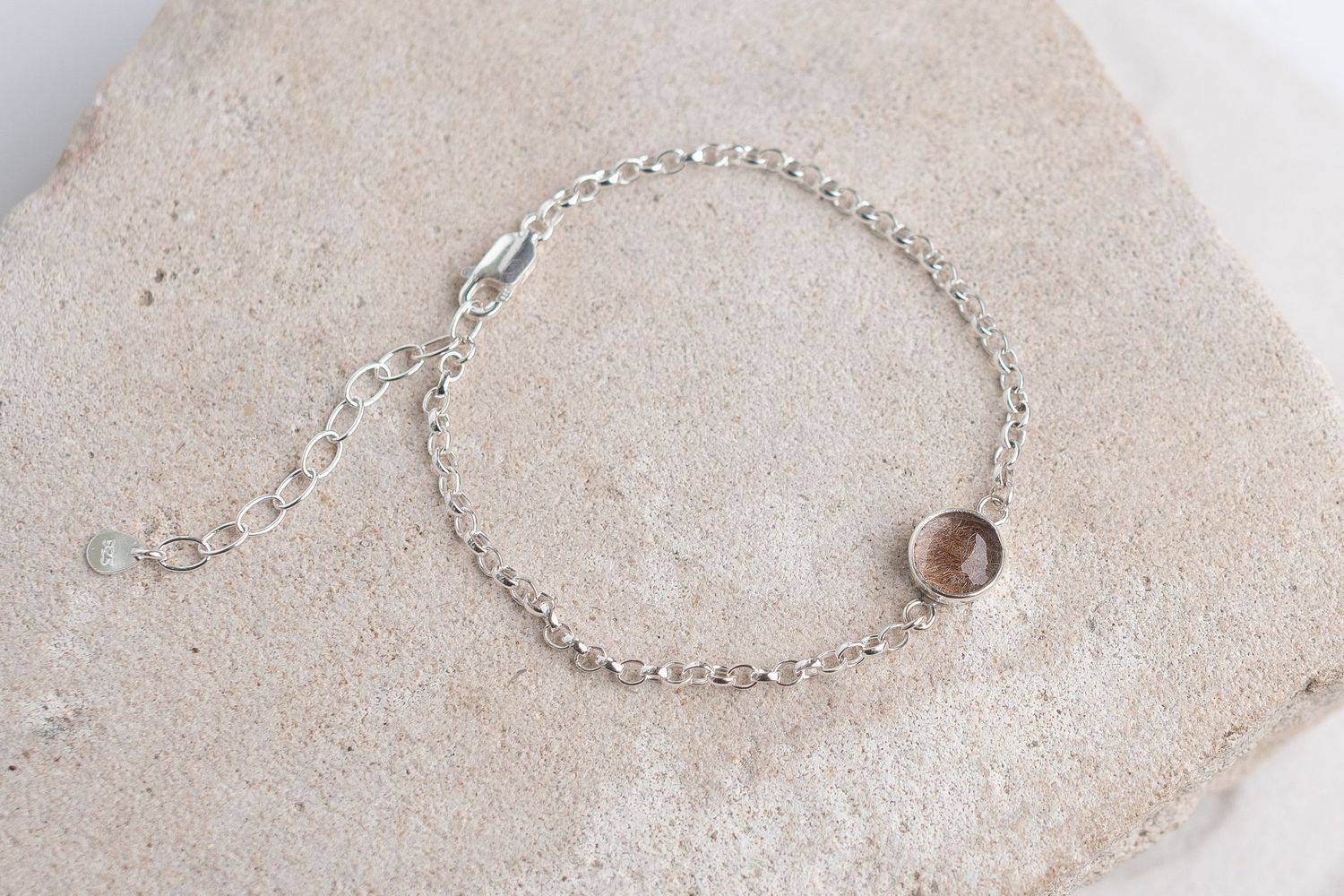 Minimalist silver chain bracelet with small circular gemstone charm displayed on a sandy stone surface.