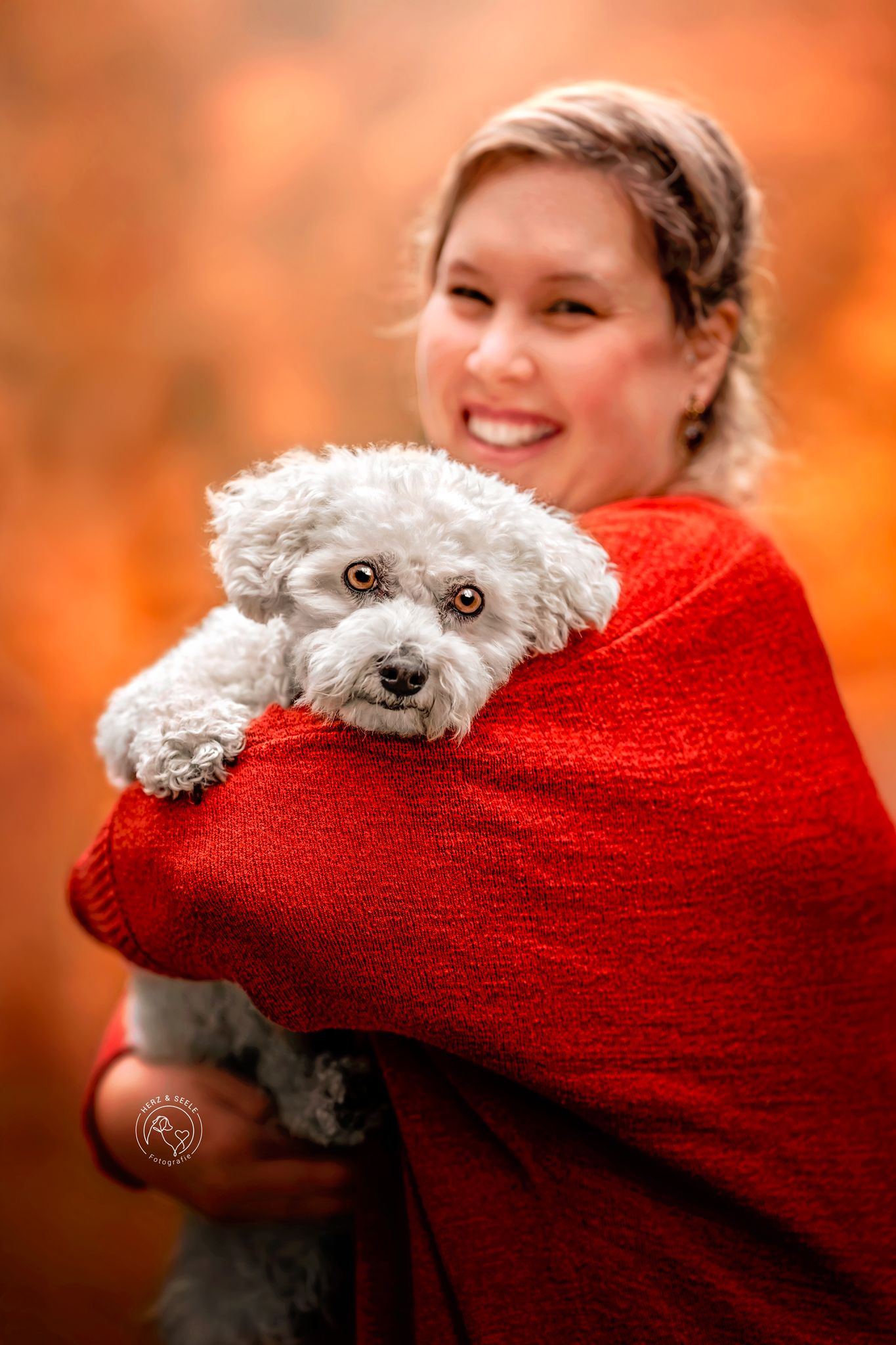 Woman in red sweater holding a fluffy white Bichon Frise dog against autumn orange foliage background.