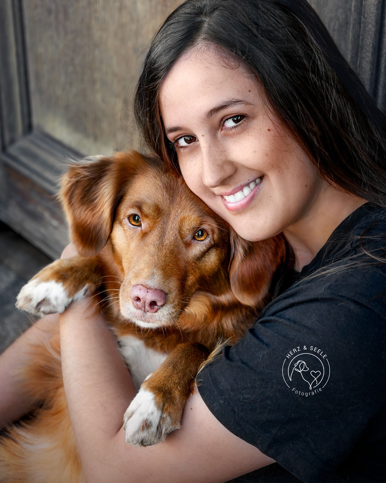 A smiling young woman with dark hair cuddles a Nova Scotia Duck Tolling Retriever dog.