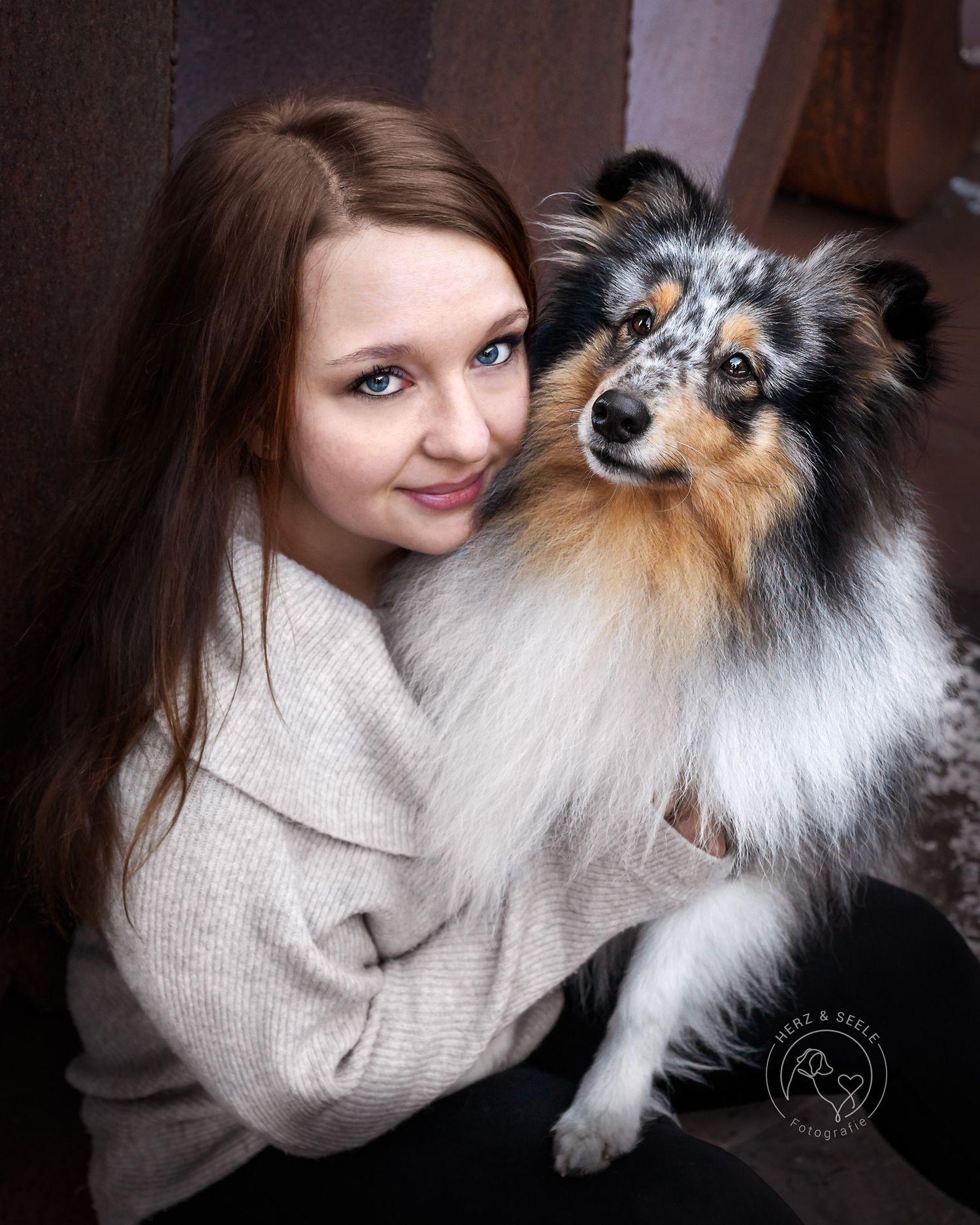 A smiling woman with brown hair poses closely with her blue merle Shetland Sheepdog indoors.