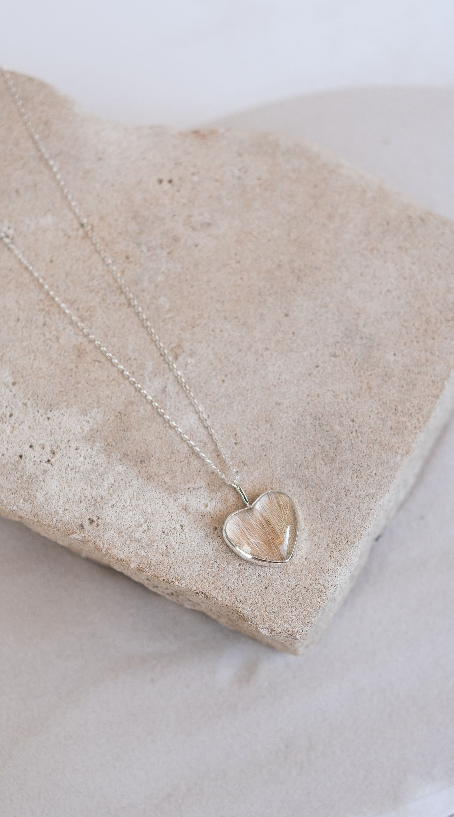 Silver heart shaped pendant necklace with rutilated quartz stone displayed on a beige stone surface.