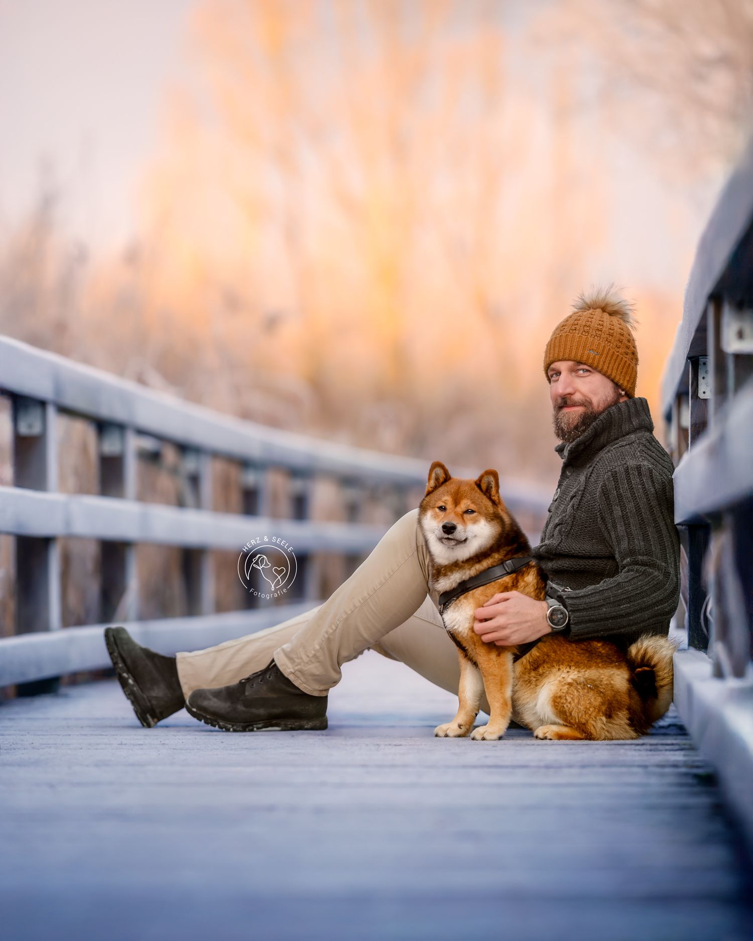 Bearded man in knit sweater and orange beanie sits with Shiba Inu dog on snowy wooden bridge.