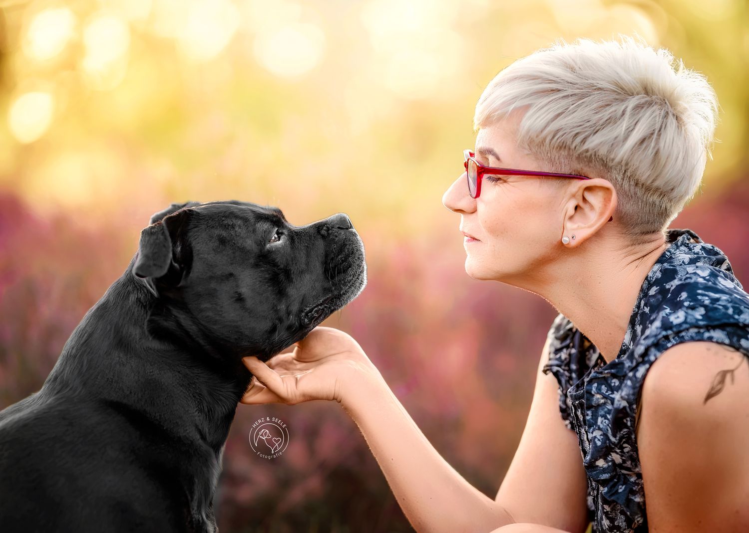 A woman with short blonde hair and red glasses gazes lovingly at a black Staffordshire Bull Terrier dog.