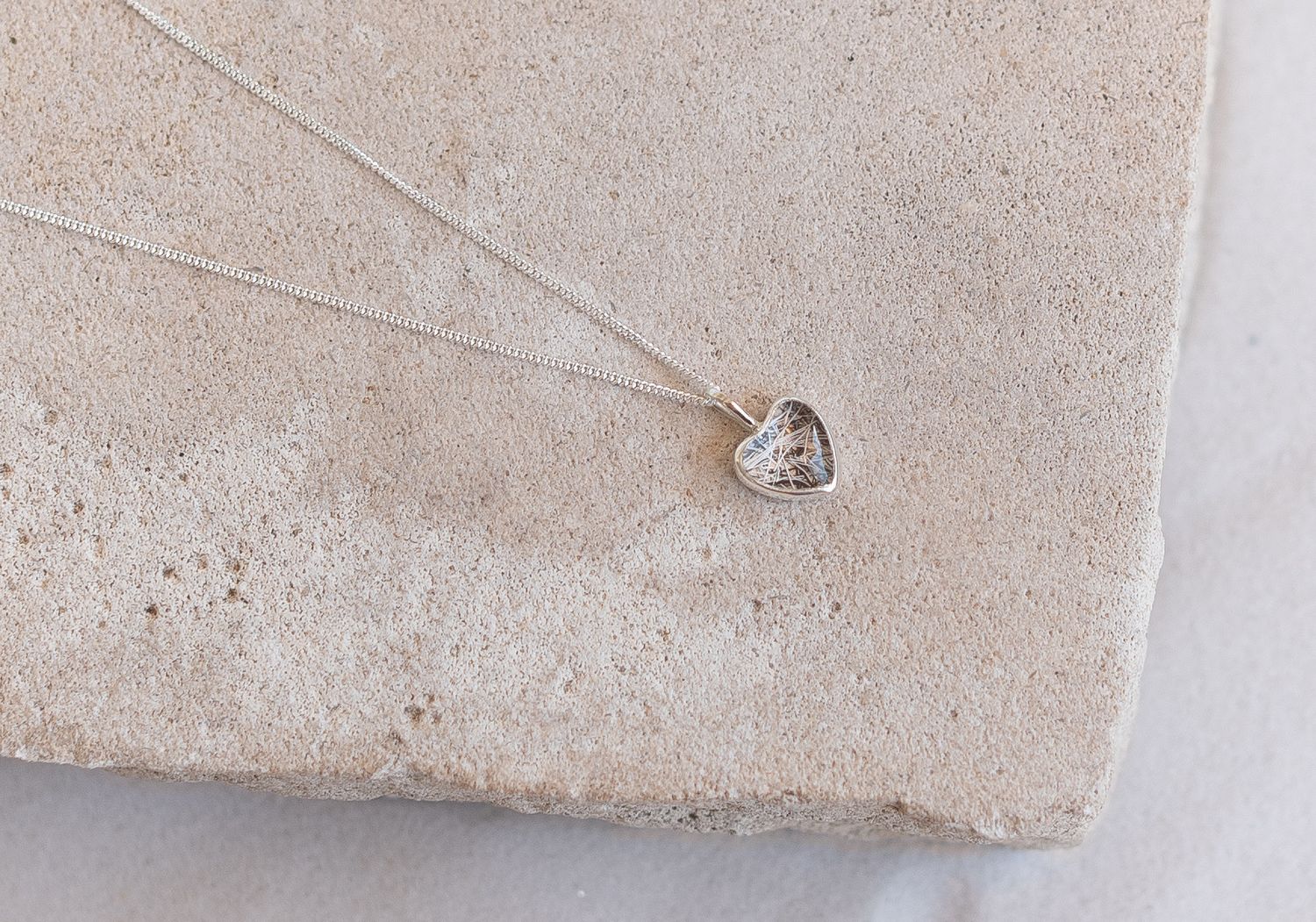 Delicate silver heart pendant necklace with textured detail displayed on a beige stone surface.