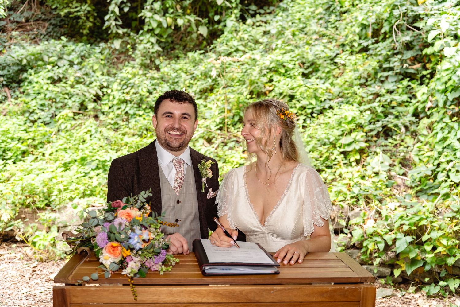 Smiling newlyweds sign wedding register at wooden table surrounded by lush green garden backdrop.