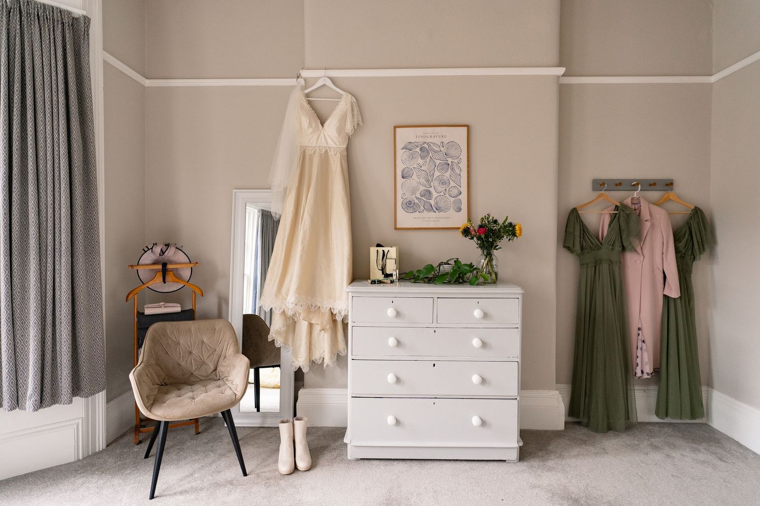 Bridal suite with white dresser, hanging wedding dress, green bridesmaid gowns, mirror, and chair in neutral-toned room.