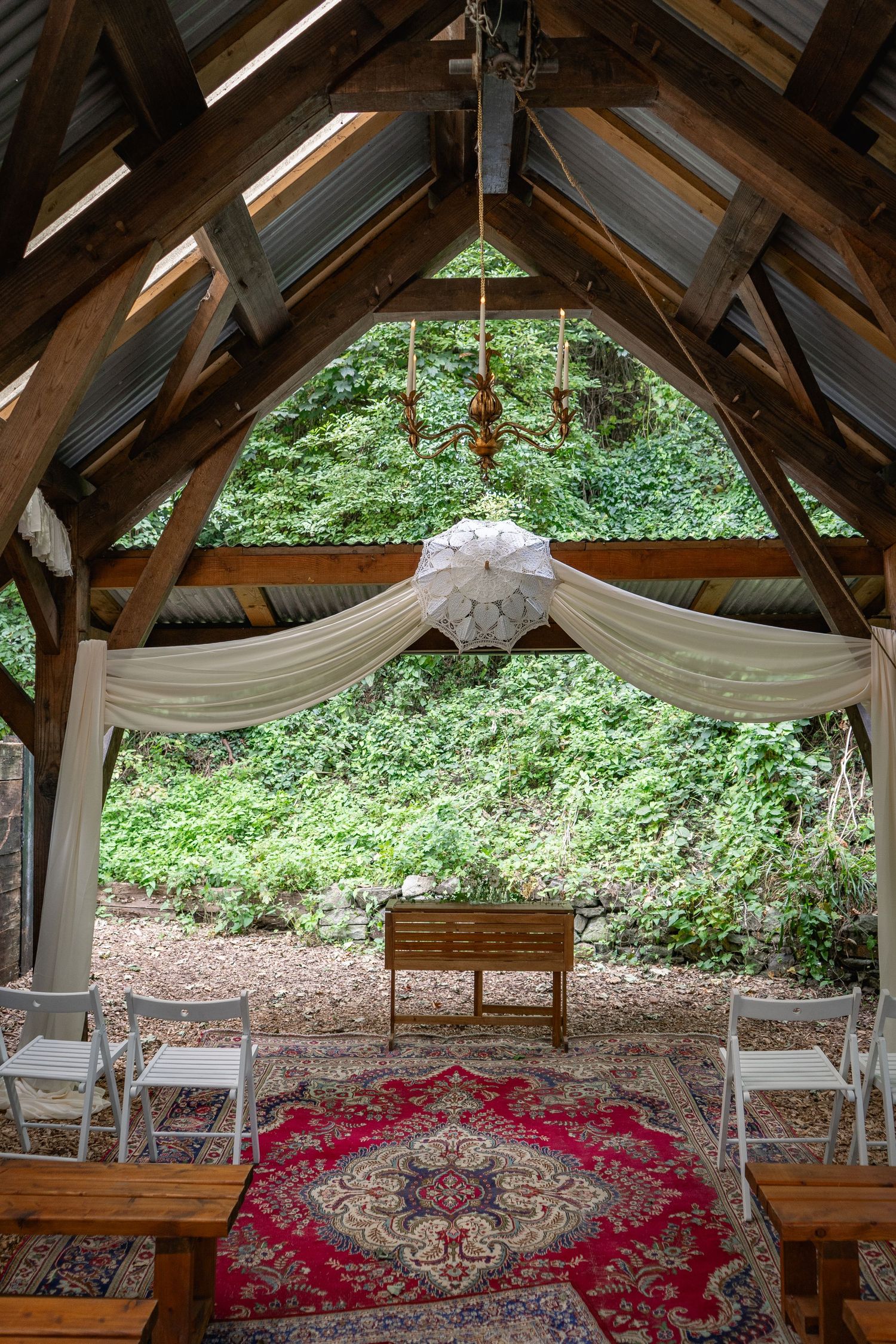 Rustic outdoor wedding chapel with white chairs, red Persian rug, draped fabric, and lush green forest backdrop.