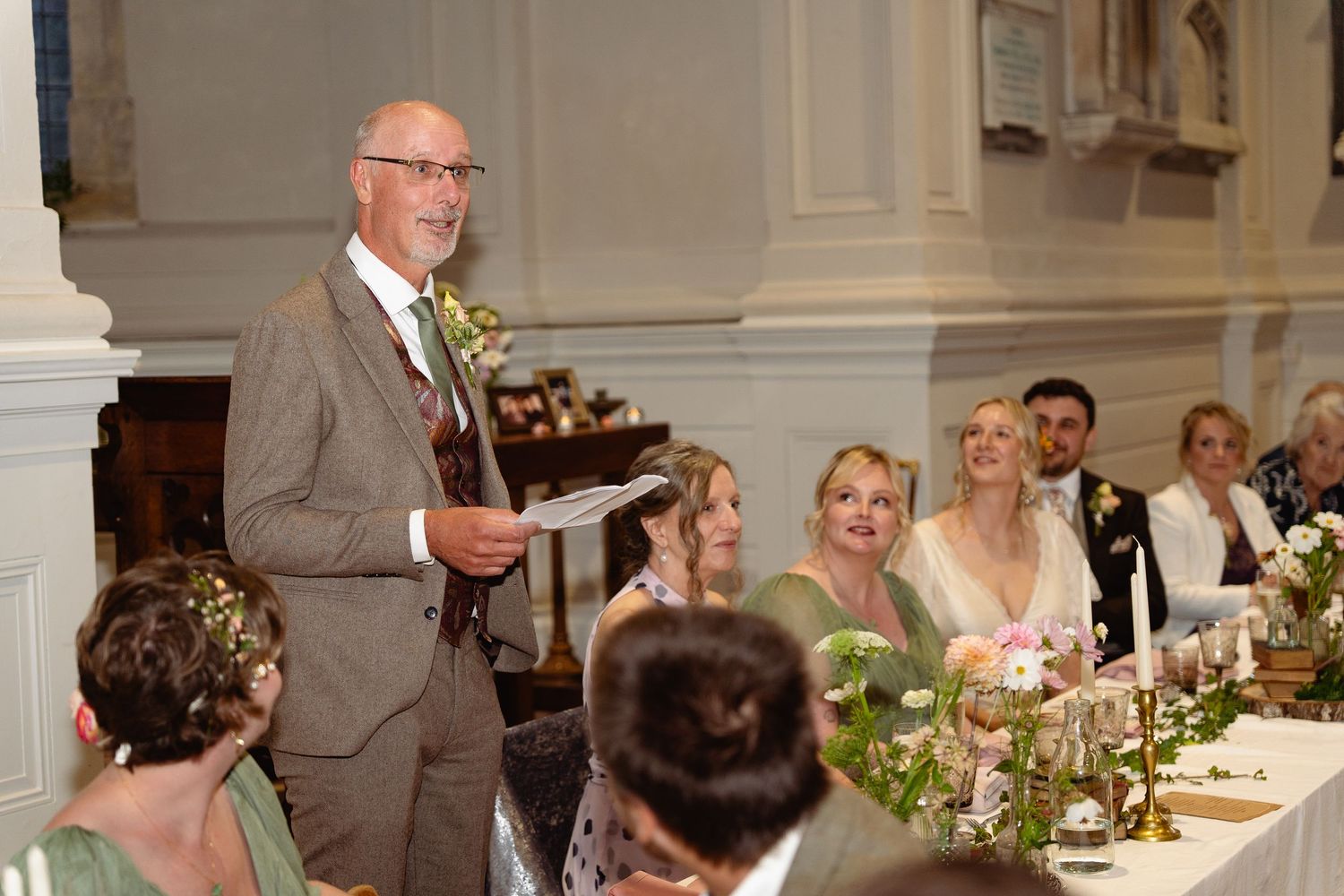 Elegant wedding speech by suited man at a floral-decorated reception with guests in a grand venue.
