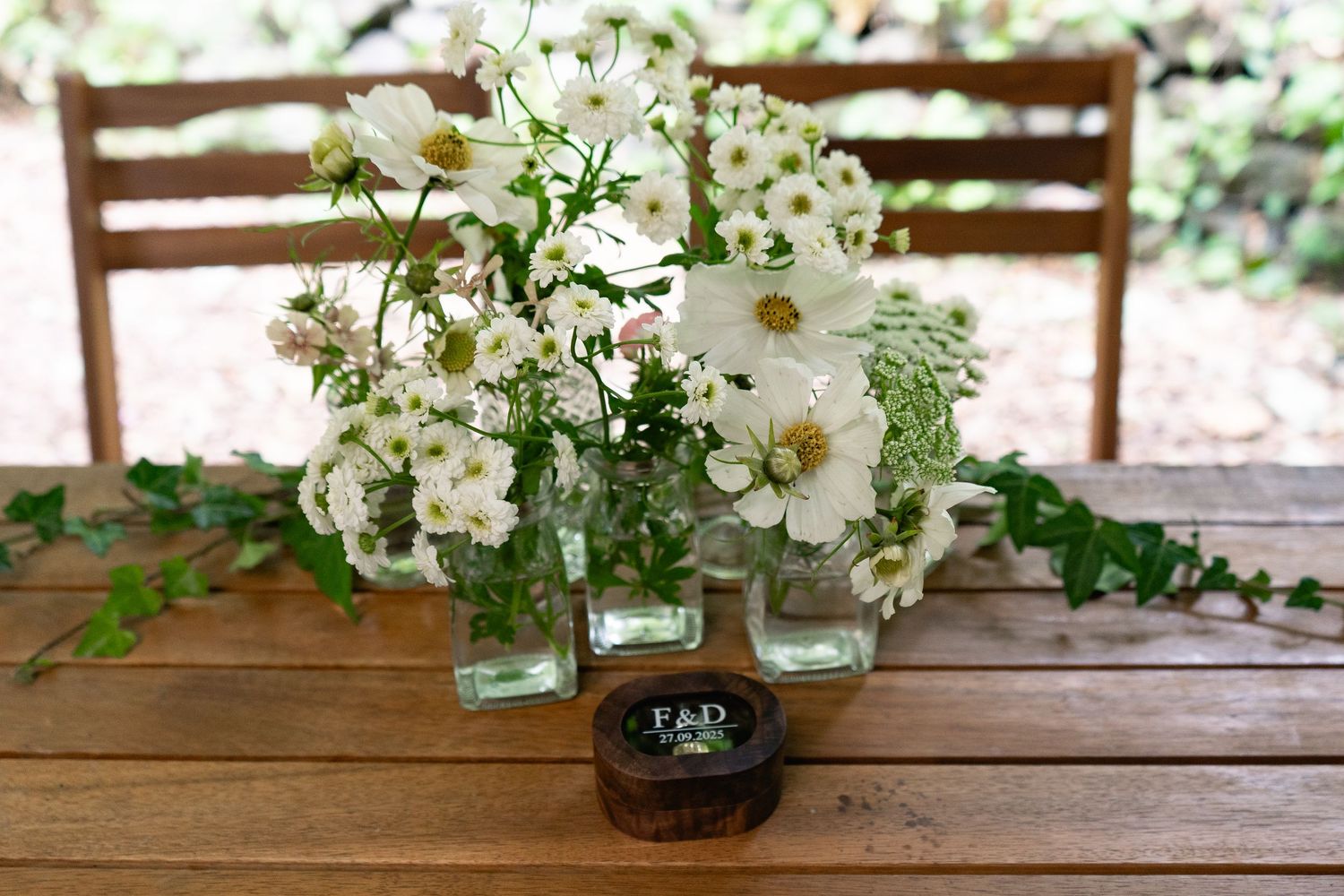 White cosmos flowers and greenery arranged in glass vases beside a small wooden ring box on a rustic wooden table.