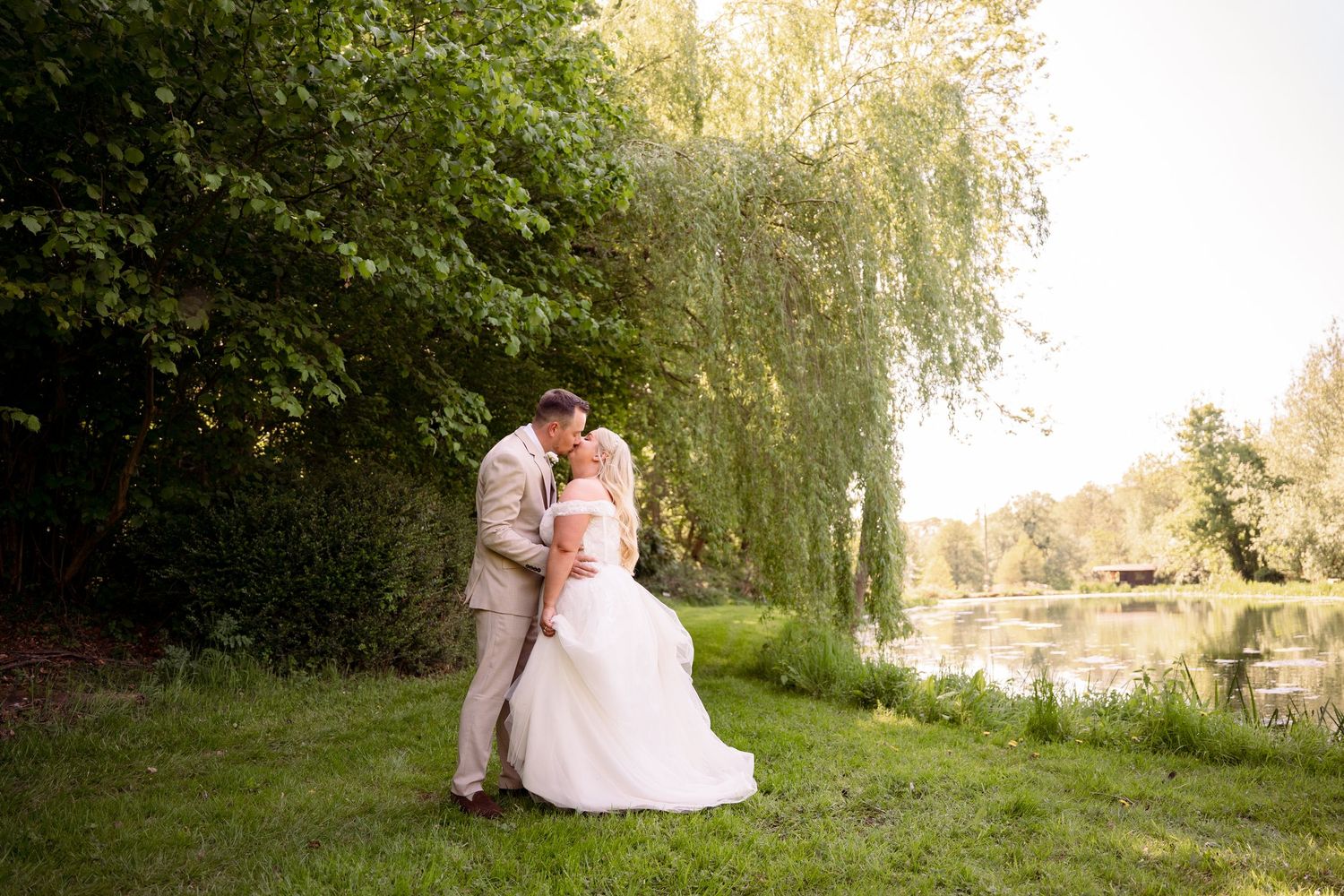 Bride and groom sharing a romantic kiss by a serene pond in Oxfordshire surrounded by lush green weeping willow trees.