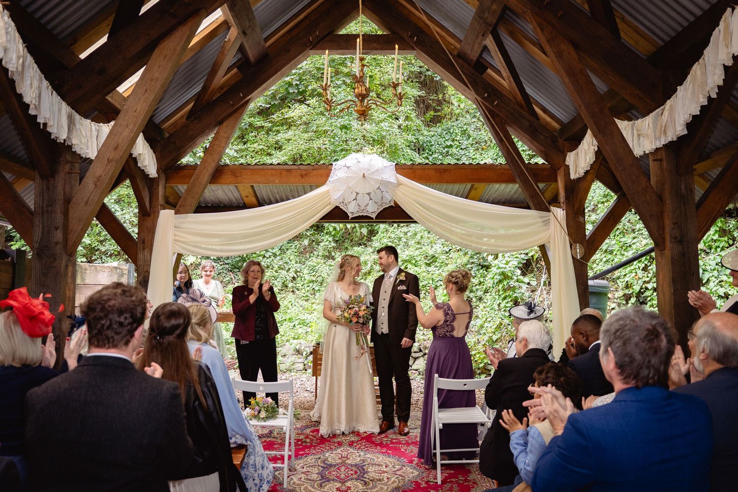 Bride and groom stand at altar during outdoor woodland wedding ceremony with seated guests watching.