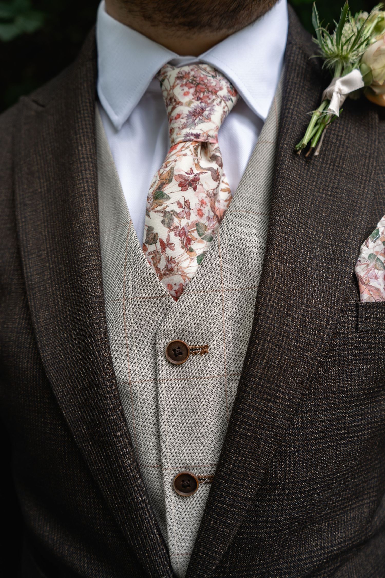 Close-up of groom's brown tweed suit with floral tie, boutonniere, and matching pocket square detail.