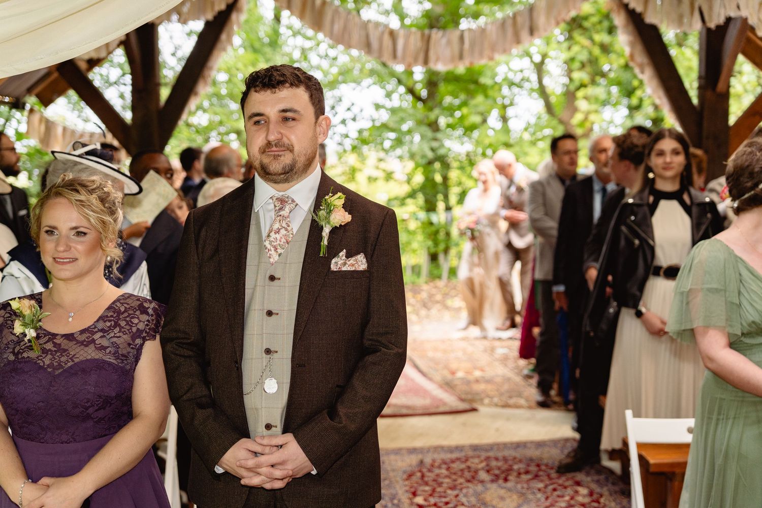 Groom in brown suit with floral tie stands at outdoor woodland wedding ceremony aisle with guests seated behind.