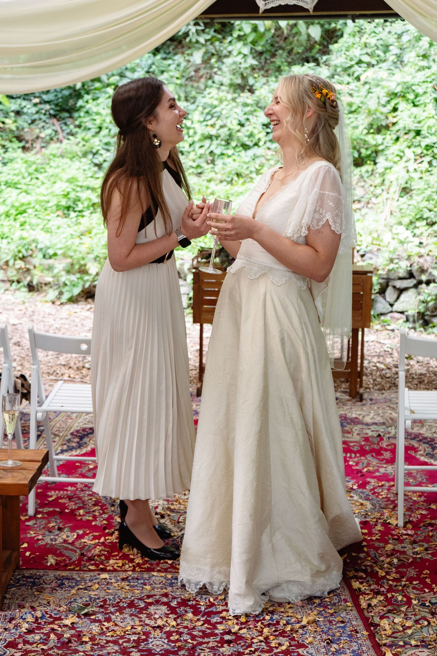 Two women in elegant dresses laugh together at an outdoor wedding ceremony on a red rug.