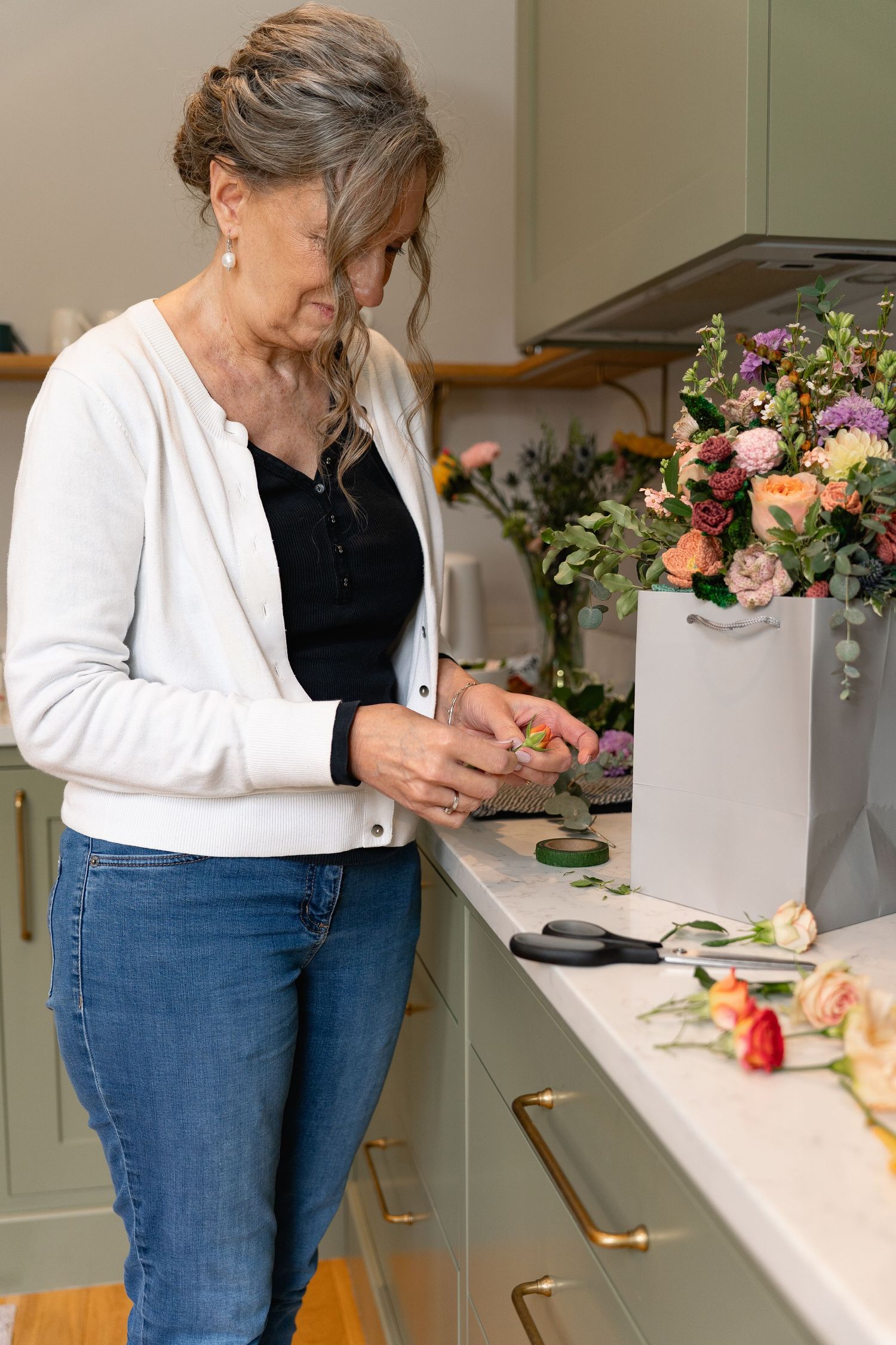 Senior woman arranging fresh flowers at a green kitchen counter with roses and bouquets.