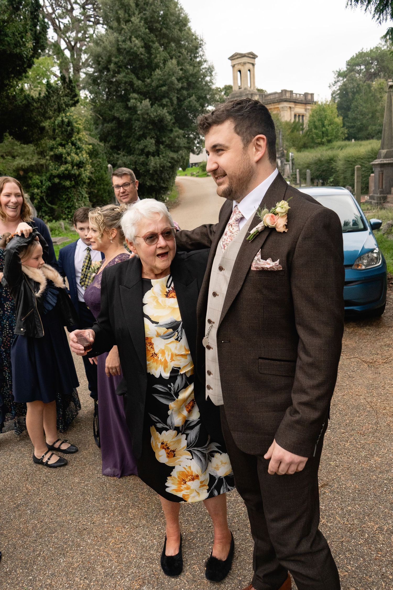 Groom in brown suit with boutonniere poses with elderly woman in floral dress at outdoor wedding venue.