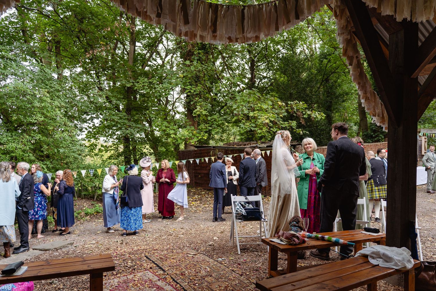 Outdoor woodland wedding reception with guests mingling under a rustic canopy adorned with bunting and surrounded by lush green trees.