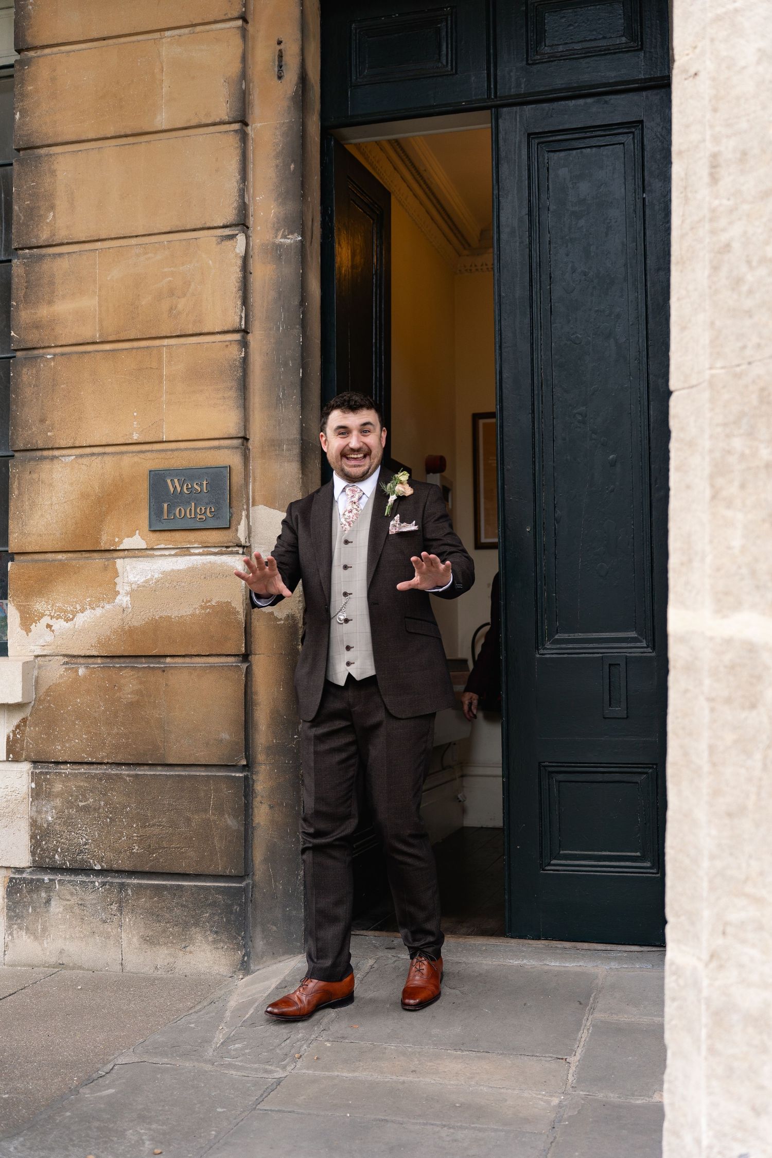 Smiling groom in brown suit with boutonniere stands at West Lodge entrance, welcoming guests on his wedding day.