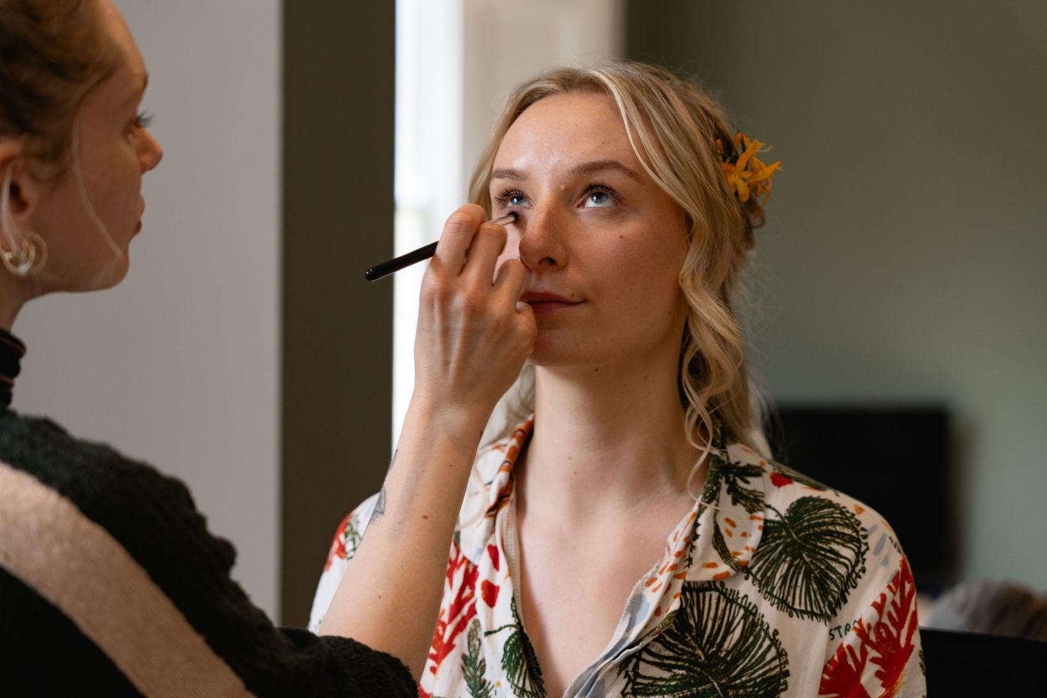 A makeup artist applying eye makeup to a blonde bride wearing a floral robe before her wedding day.