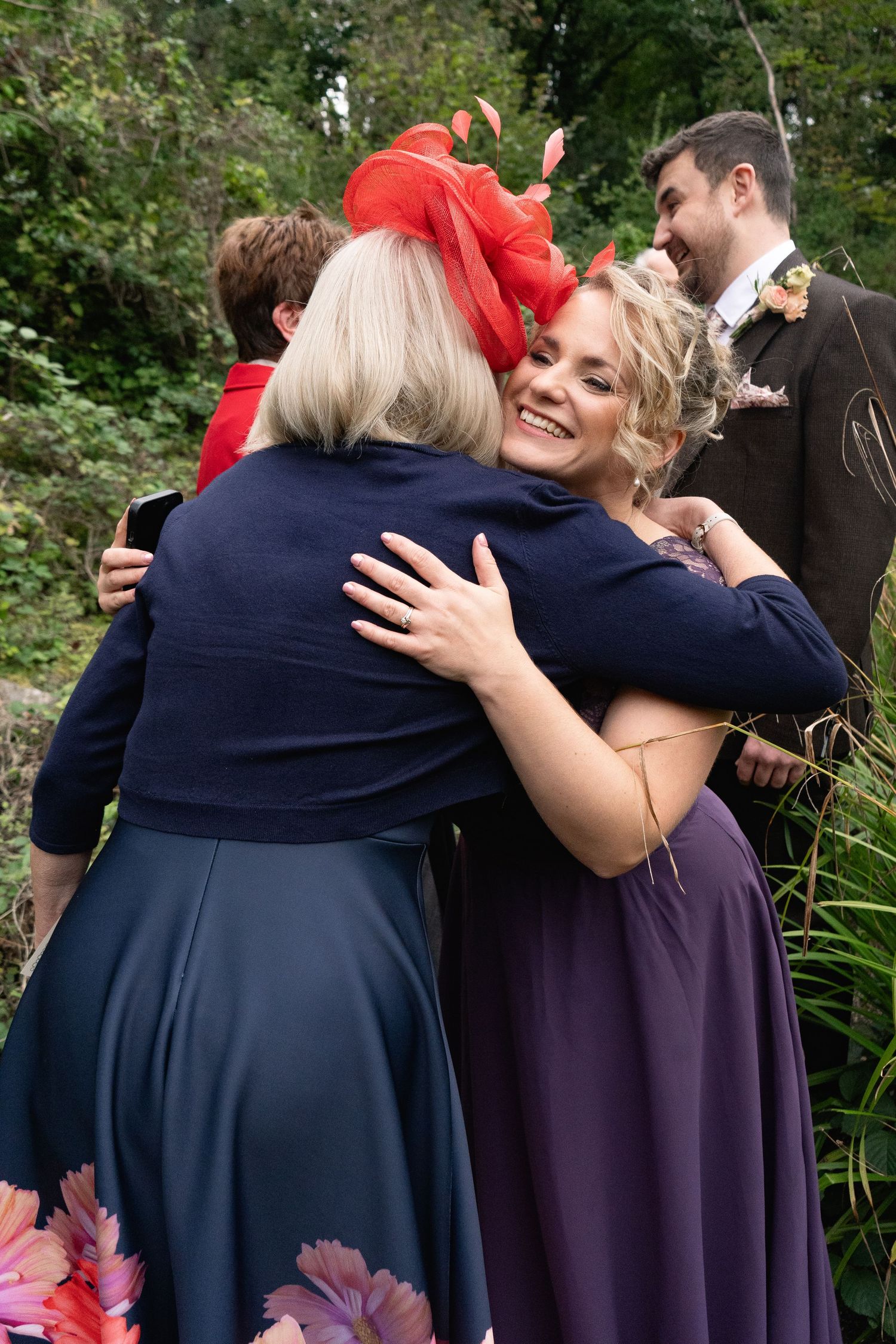 Bride in purple dress hugging guest in navy at outdoor wedding celebration with groom visible in background.