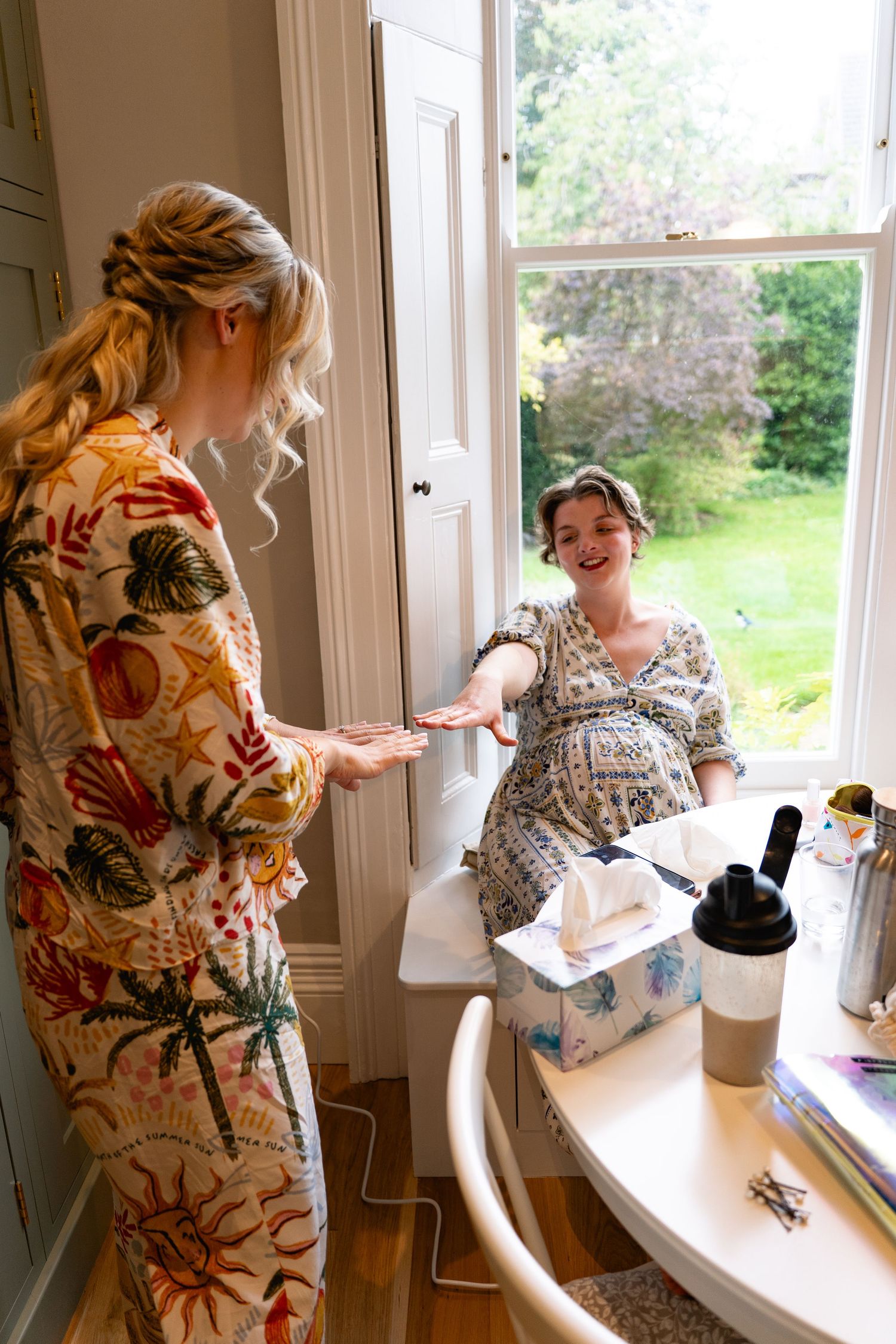 Pregnant woman in floral dress receives item from blonde woman in tropical robe near bright window during wedding preparations.