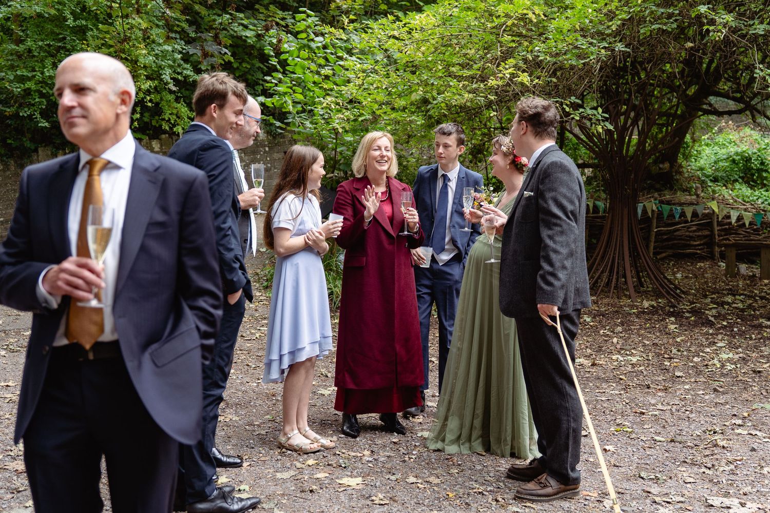 Elegantly dressed wedding guests socializing outdoors in a lush garden setting, holding champagne glasses.