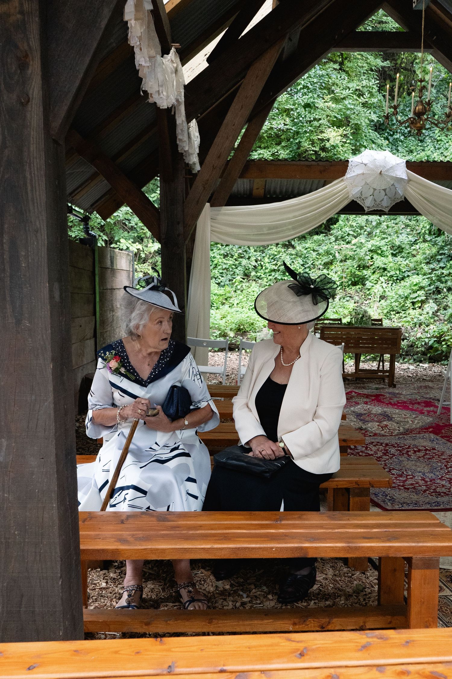 Two elegantly dressed elderly women wearing decorative hats sit conversing on wooden benches under a rustic outdoor pavilion.