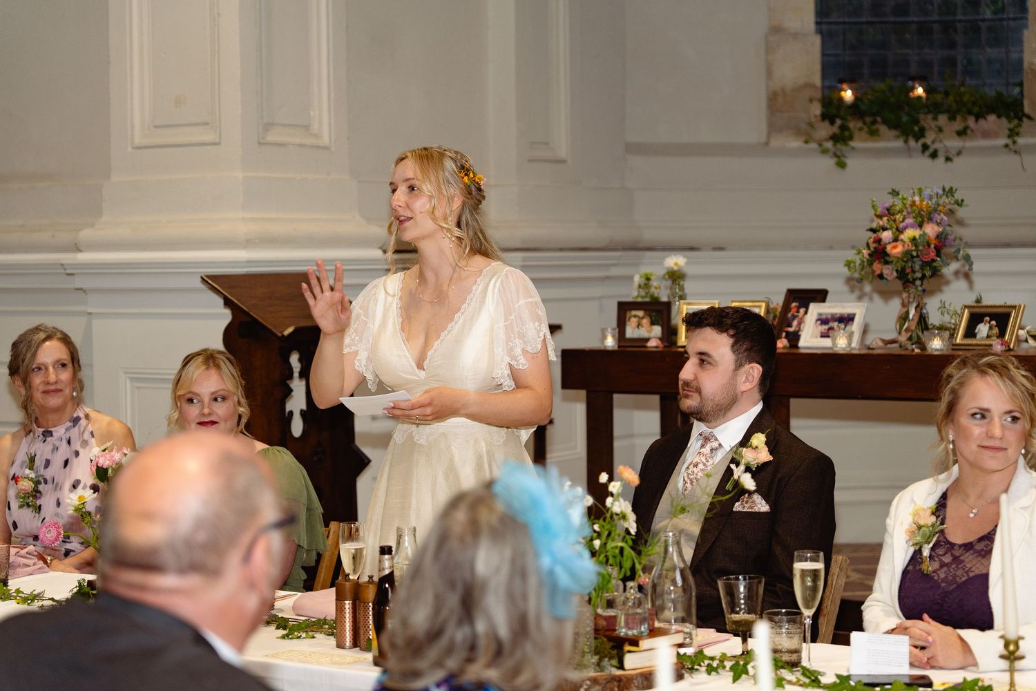 Bride giving a speech at wedding reception dinner table with guests seated around her.
