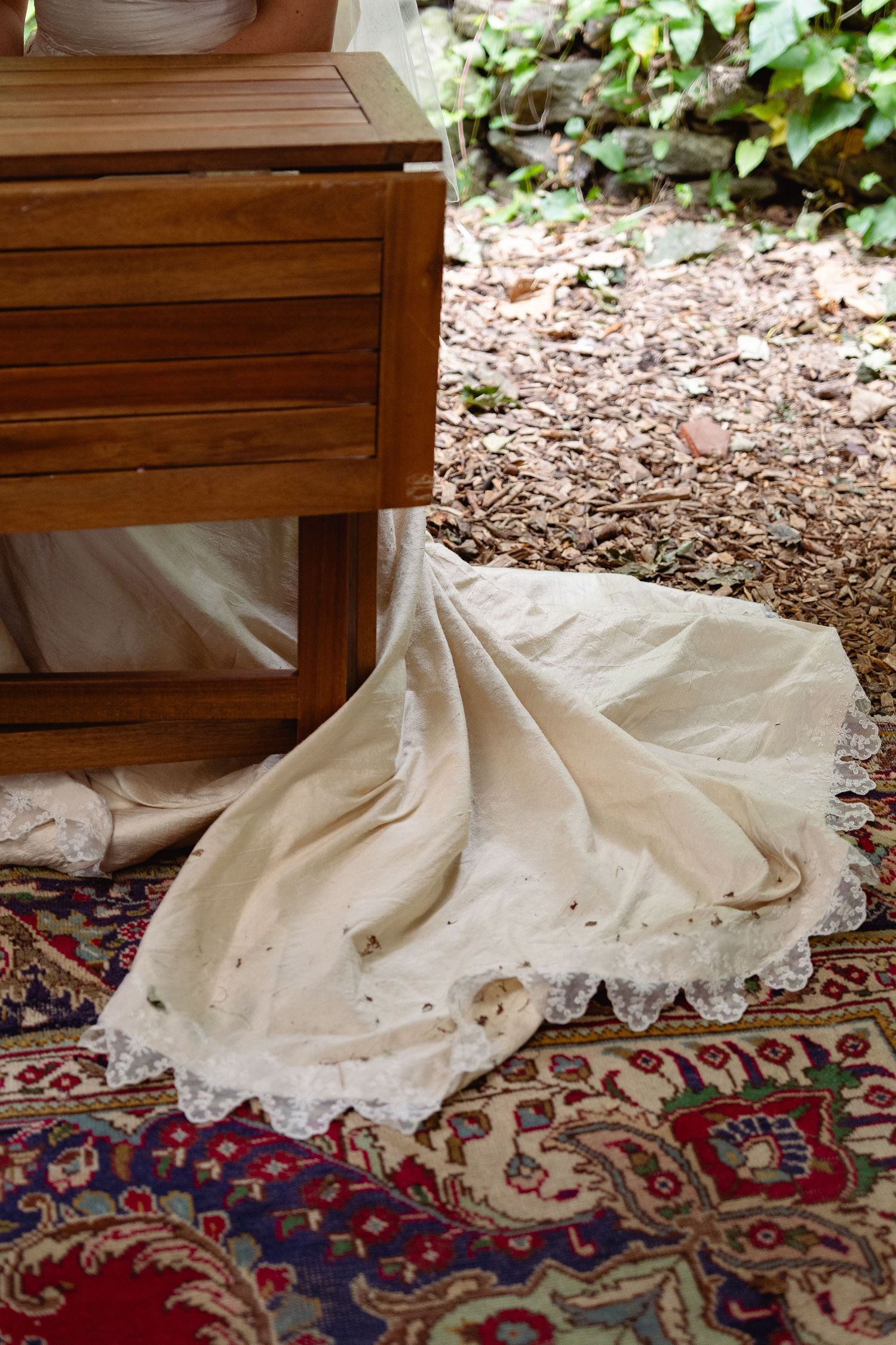 Elegant white wedding dress train with lace trim spread across a colorful Persian rug beside a wooden table outdoors.