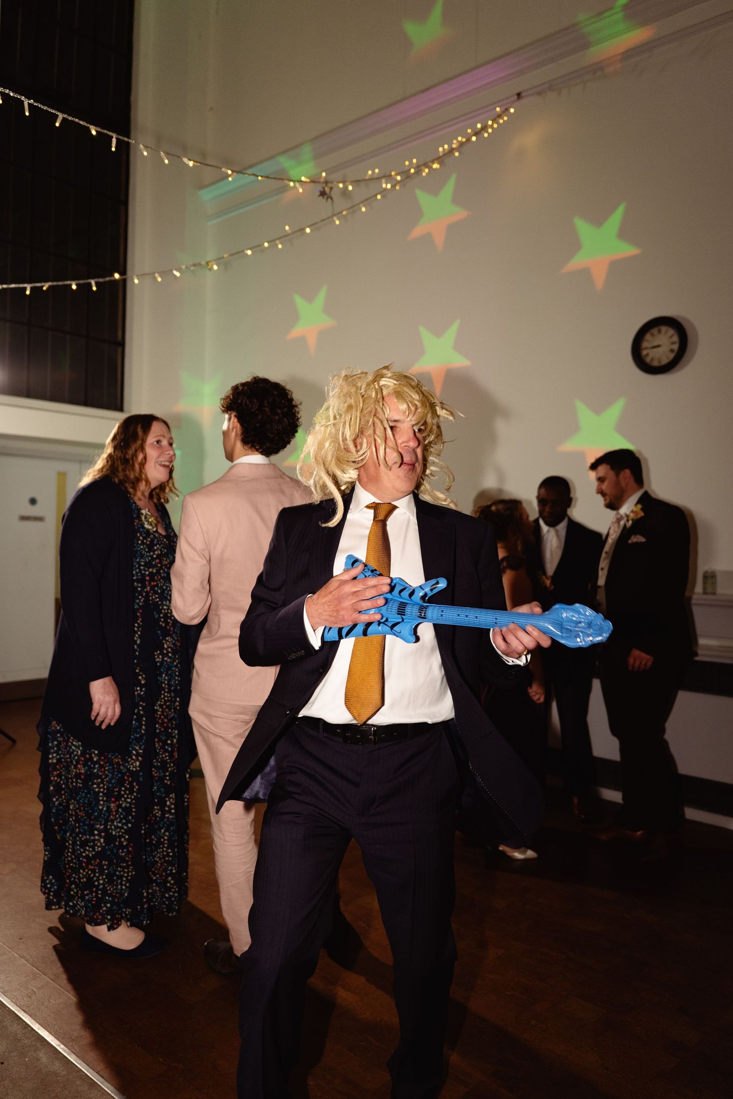 Man in suit with blonde wig dancing energetically with a blue balloon guitar at a lively wedding reception.