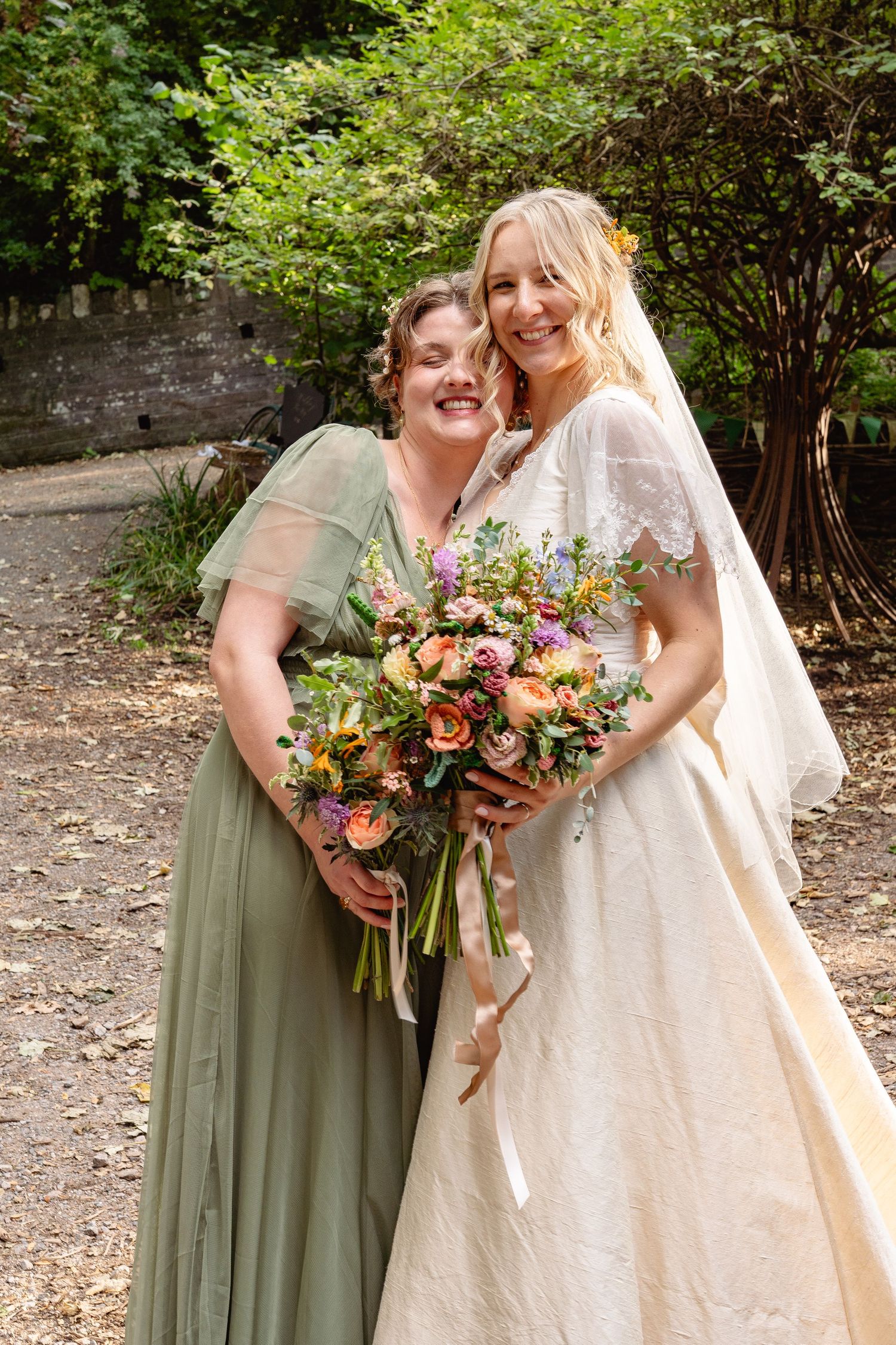 A smiling bride in a lace wedding gown and veil holds a bouquet beside her bridesmaid in a sage green dress.