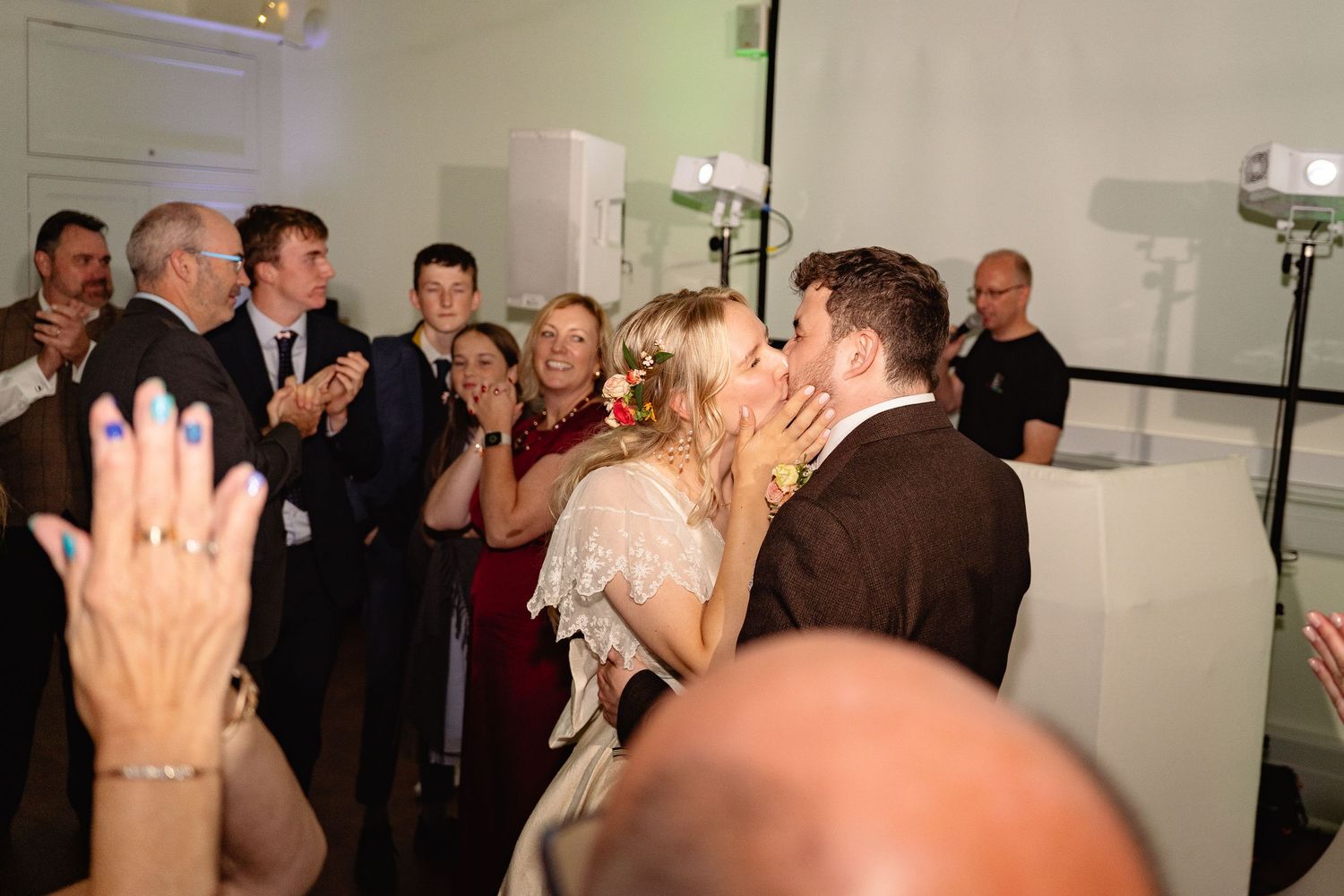 Newlyweds share a kiss surrounded by cheering guests at a wedding reception celebration.
