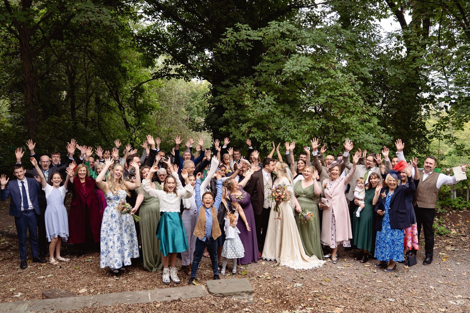 Large wedding group photo with guests waving hands outdoors, surrounded by lush green trees.