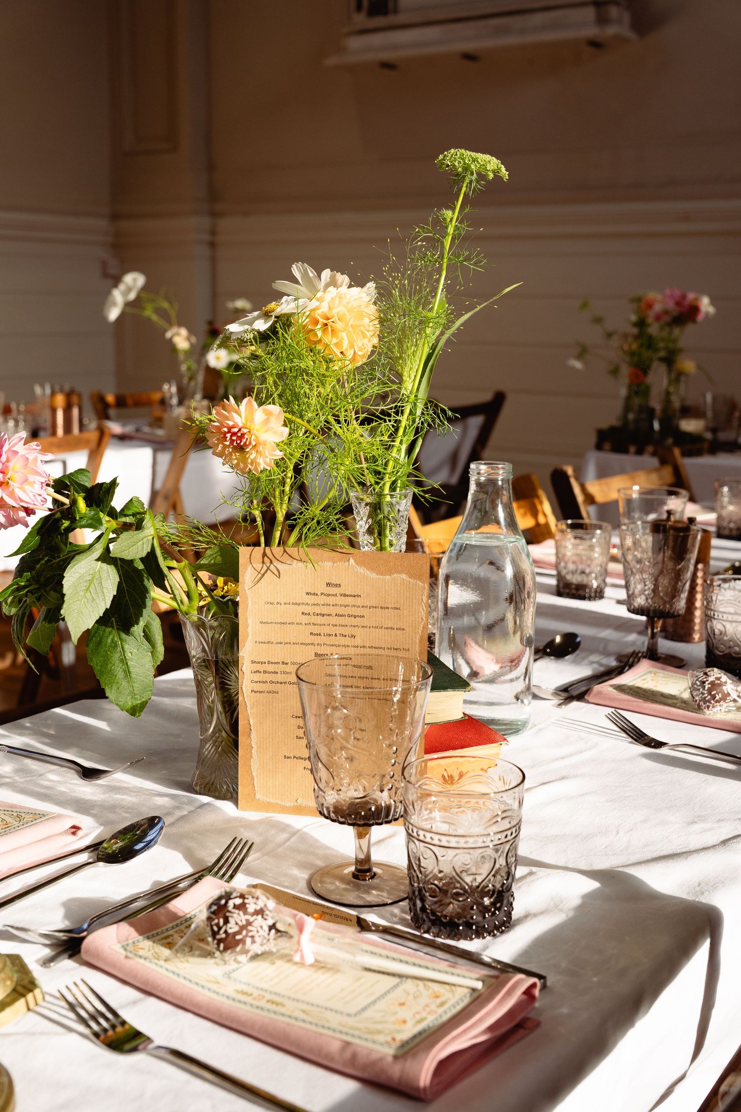 Elegant wedding reception table setting with wildflower centerpiece, crystal glassware, pink napkins, and menu cards.