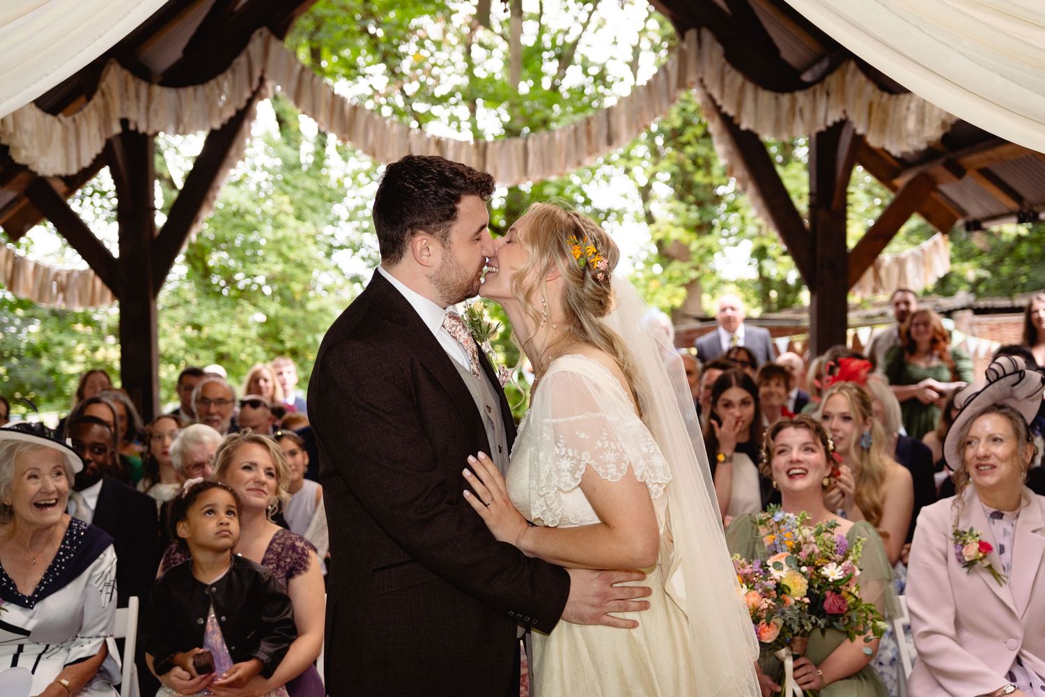 Bride and groom share their first kiss at an outdoor wedding ceremony with guests applauding.