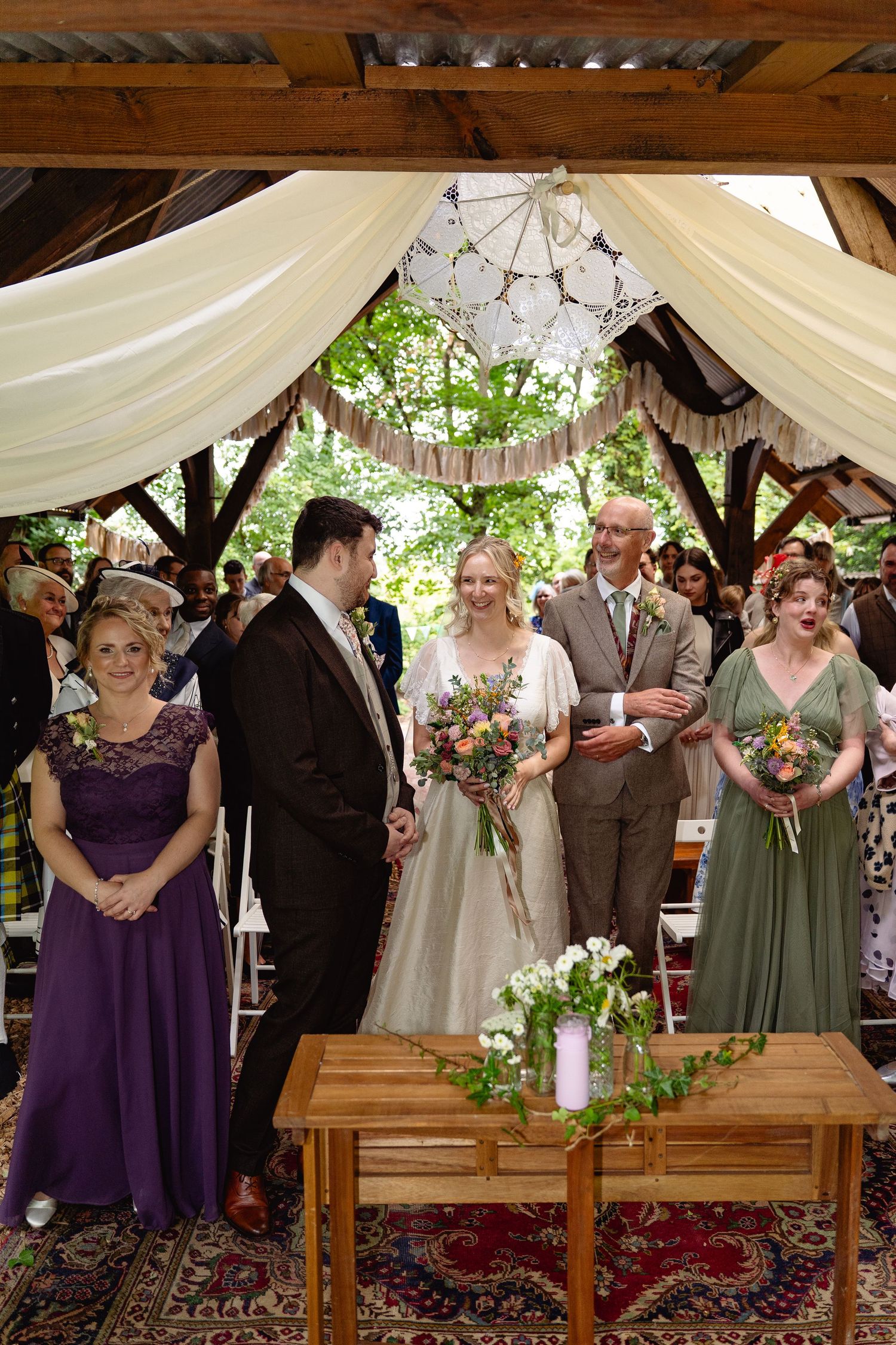 Wedding party stands at outdoor altar under draped fabric canopy with bride, groom, and attendants in sage green and purple.