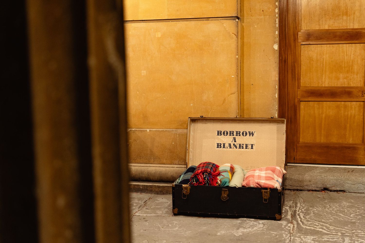 Open vintage suitcase labeled Borrow a Blanket filled with colorful blankets, placed on floor near wooden door.