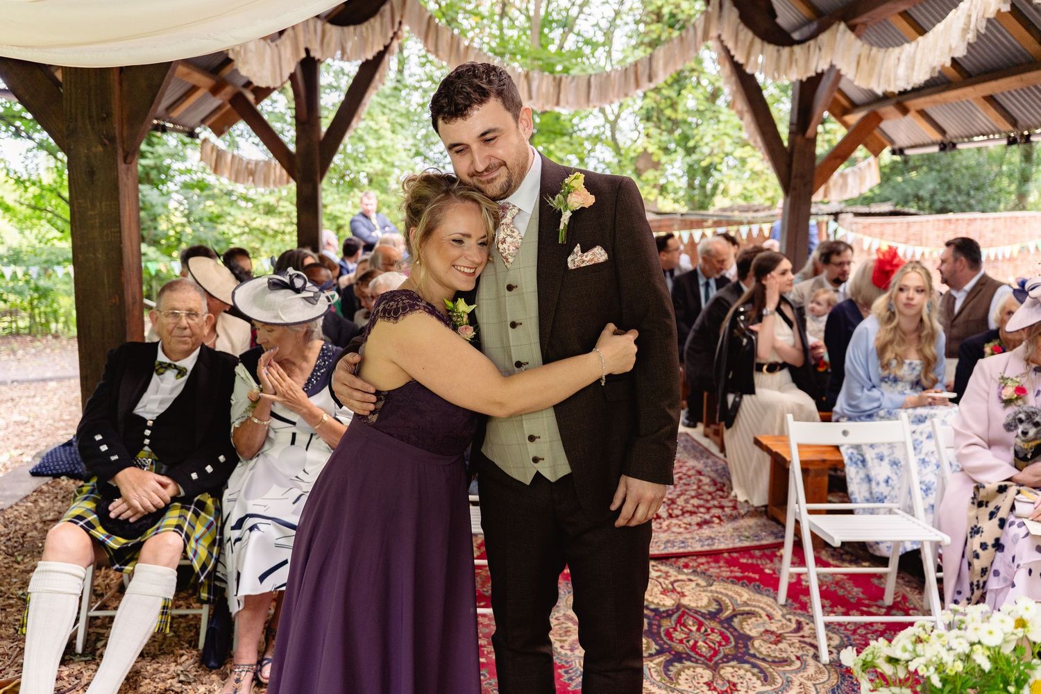 Young couple in wedding attire pose together smiling under a decorated woodland gazebo during a ceremony.