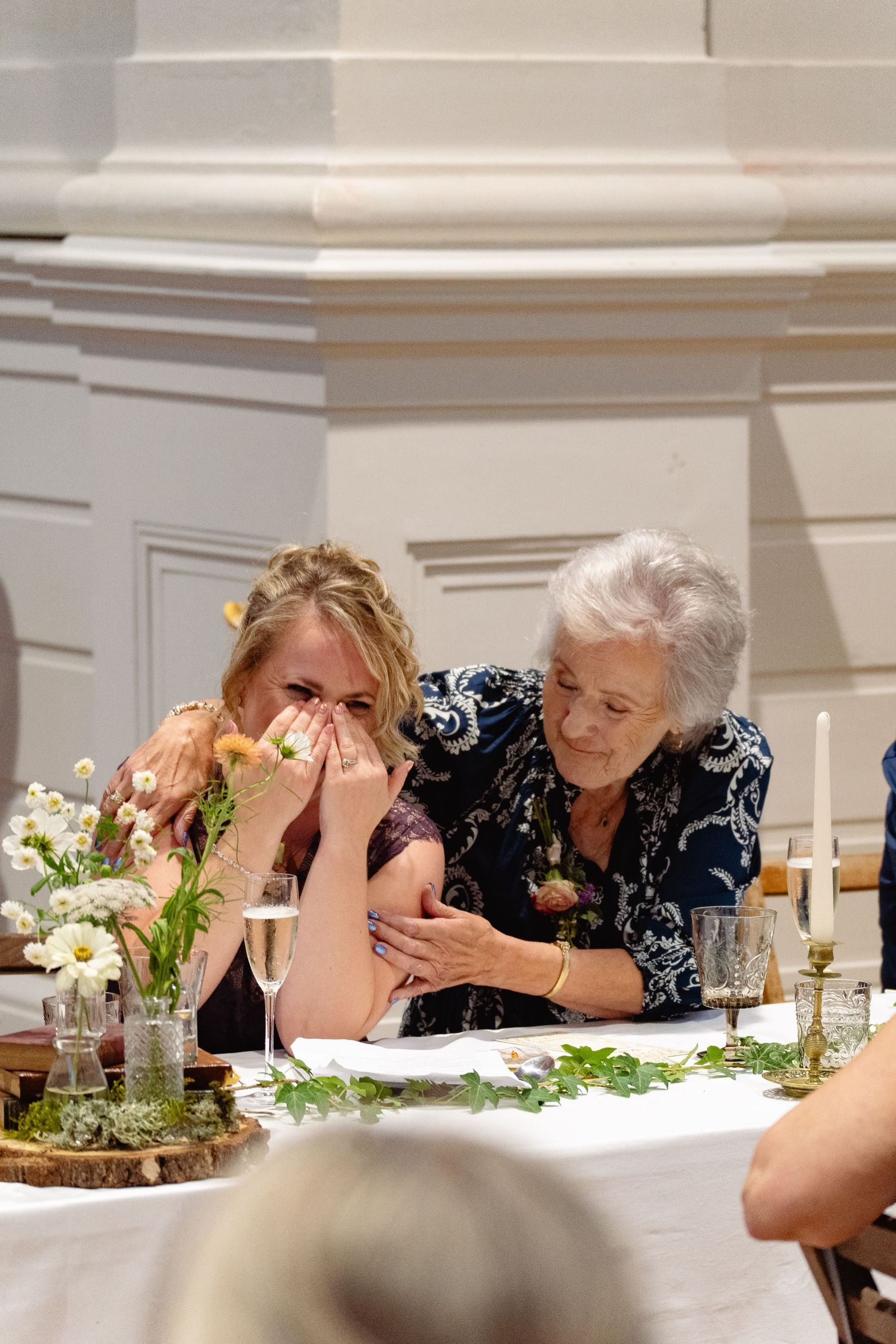 Emotional wedding guest crying while elderly woman comforts her at floral-decorated reception table in historic venue.
