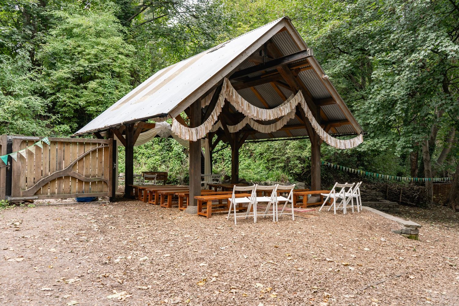 Rustic outdoor wedding pavilion with wooden benches, white chairs, and draped fabric decorations surrounded by lush trees.